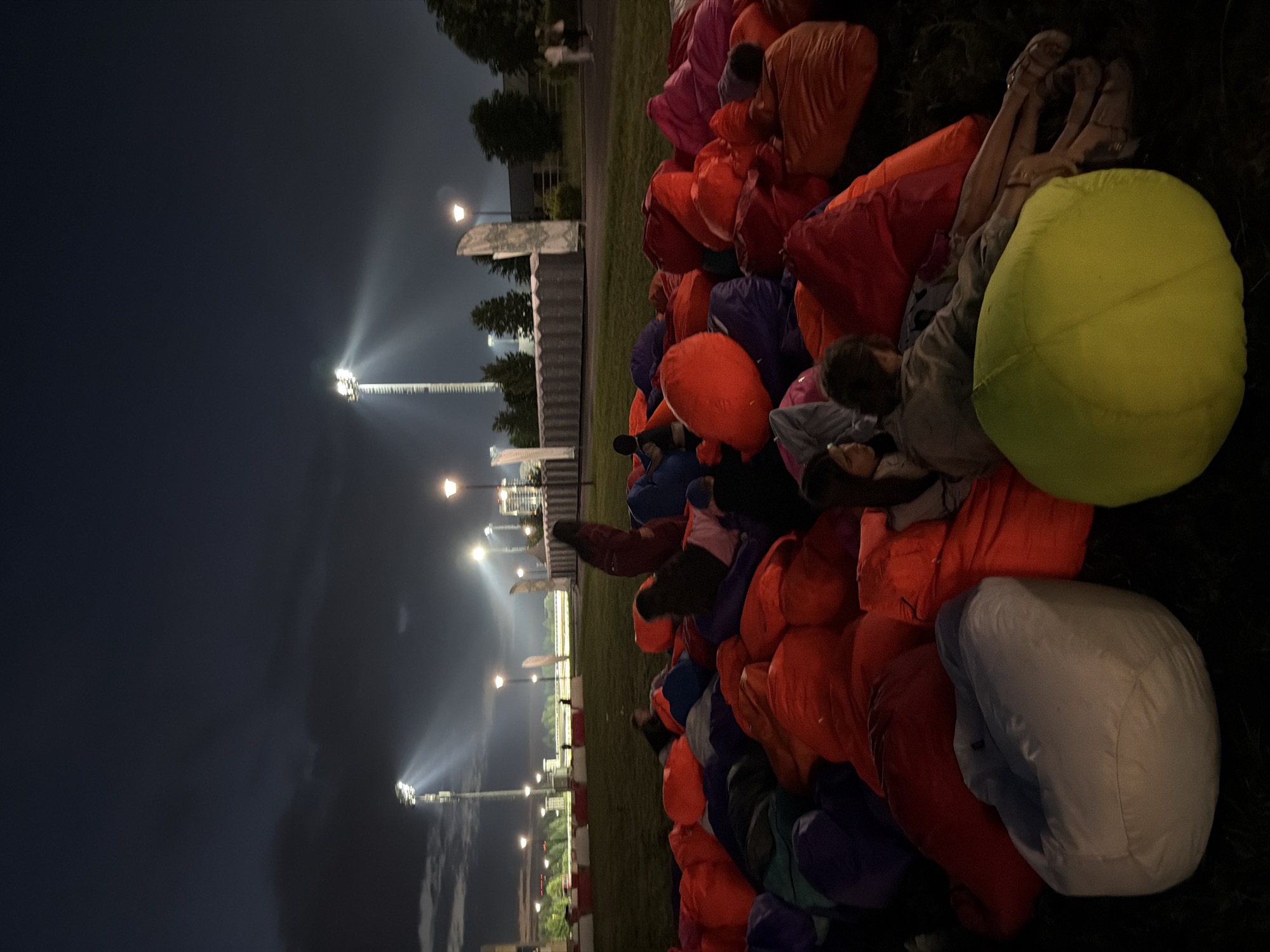 At dusk in an outdoor urban park, numerous people are gathered on a grassy area, sitting on a large collection of colorful beanbag chairs. The sky is dark blue with scattered clouds. Tall stadium lights are visible in the background, illuminating parts of the park with their bright beams. The scene appears to be a casual outdoor event, possibly a movie screening or a concert, with people relaxing on the beanbags. Some individuals are looking in the same direction, suggesting a shared focus on an event happening off-camera. The lighting suggests it is either early evening or late night.