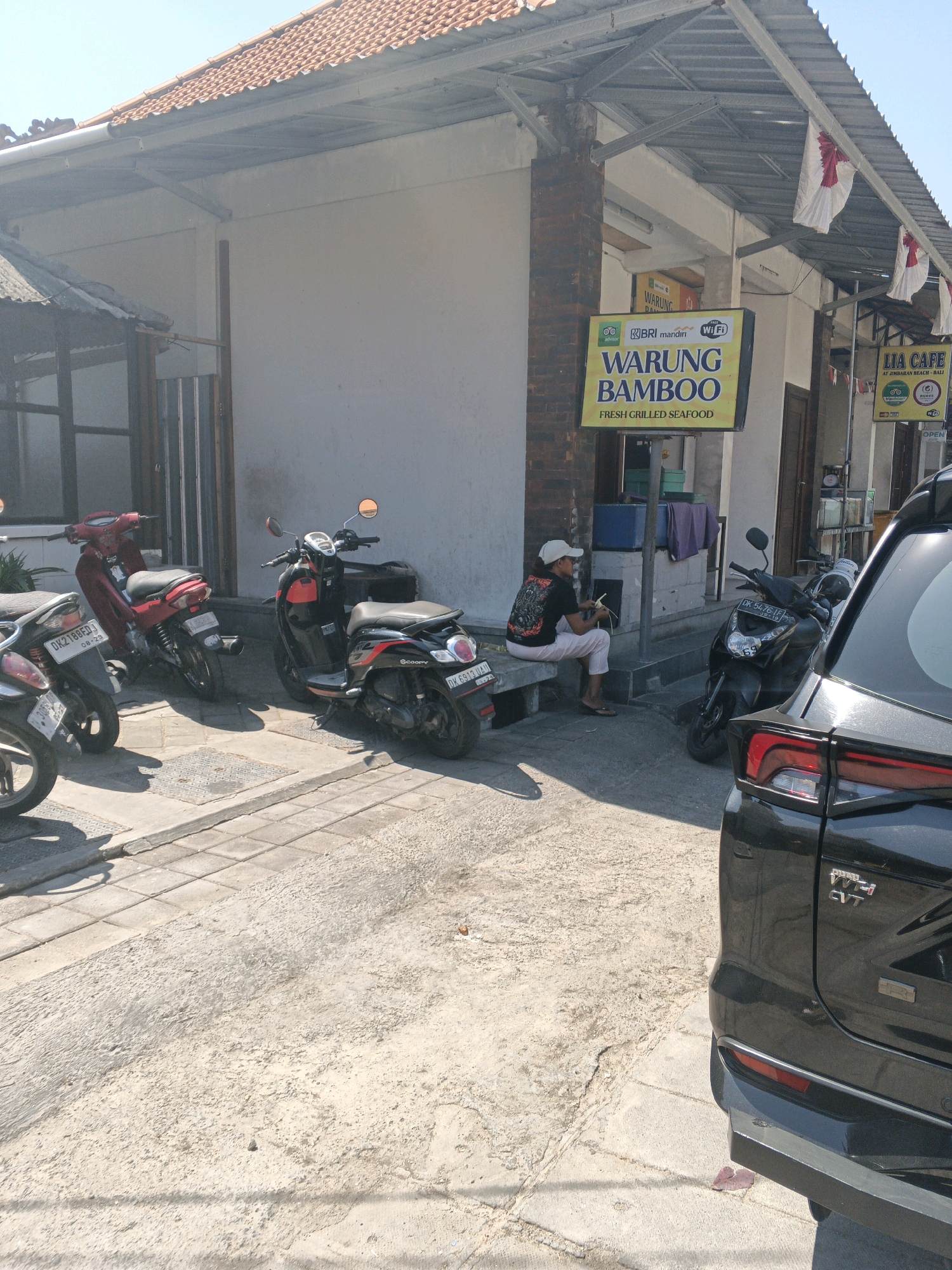 The image captures a scene in Kuta Selatan, Indonesia, likely a street in front of a commercial establishment. In the foreground, the rear of a black vehicle is visible, its taillights and rear bumper illuminated by sunlight. To the left, several scooters are parked along the sidewalk. A red scooter and a black scooter with red accents are prominently featured. Further left, the front portion of another scooter is visible.
Behind the scooters, a building with a tiled roof and white walls stands. A prominent yellow sign with red lettering reads "WARUNG BAMBOO" and "FRESH GRILLED SEAFOOD," indicating it's a food establishment. Another sign, "LIA CAFE," is visible to the right of the "WARUNG BAMBOO" sign. Several white flags with red designs are hung along the building's overhang.
A person is seated on a concrete bench in front of the building, wearing a white cap, a dark t-shirt with a graphic design, and light-colored pants. They appear to be holding something in their hands. The ground is a mix of concrete pavement and rougher, textured ground. The overall lighting suggests it is daytime with bright sunlight.