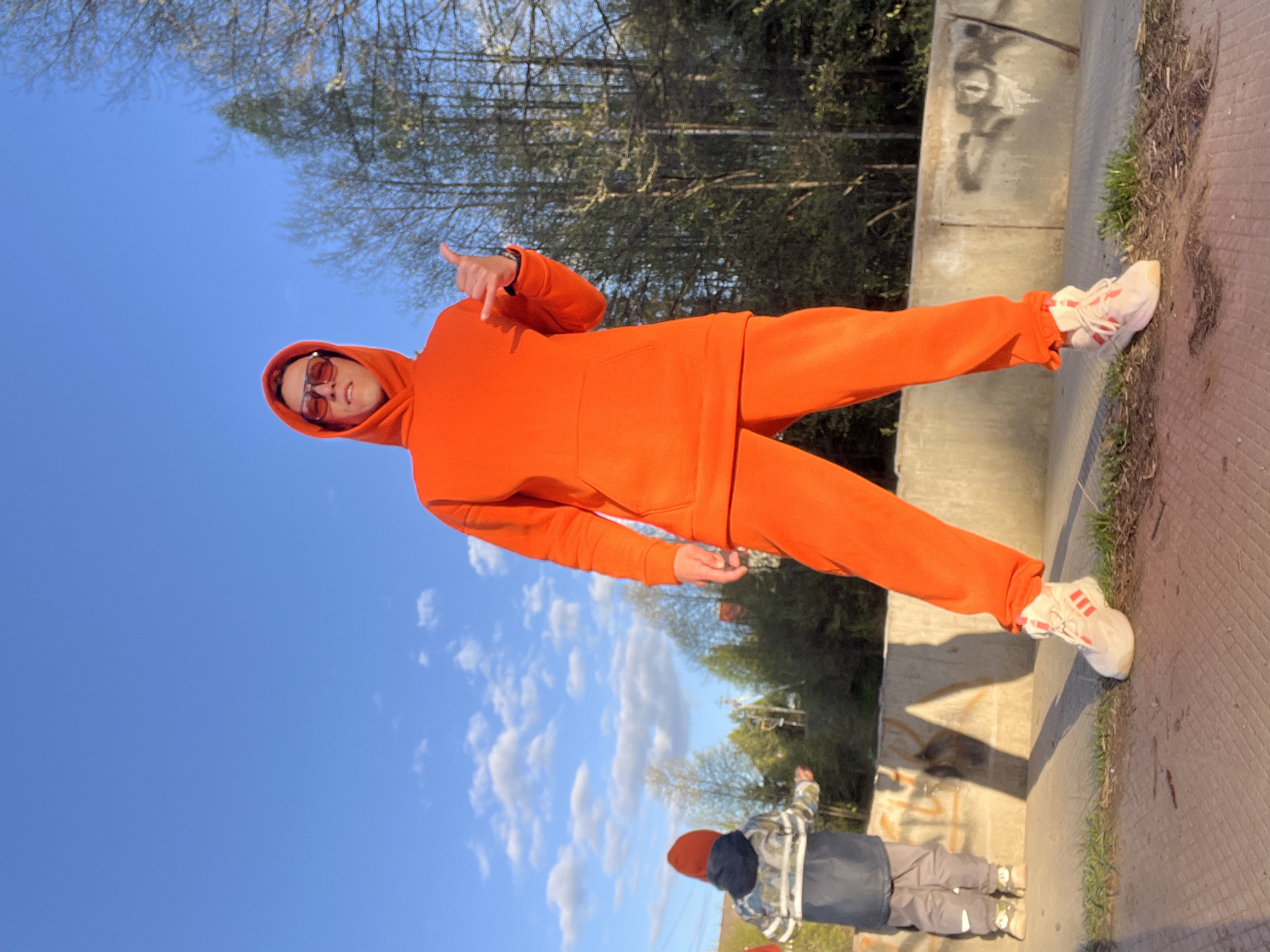 A man in an orange tracksuit and sunglasses stands on a concrete ledge, giving a thumbs-up. A child in a dark blue hat and gray jacket stands below, looking toward the trees. The location is Chaikovsky, Russia, based on available data. The background includes trees, a blue sky with clouds, and what appears to be a concrete structure with graffiti.