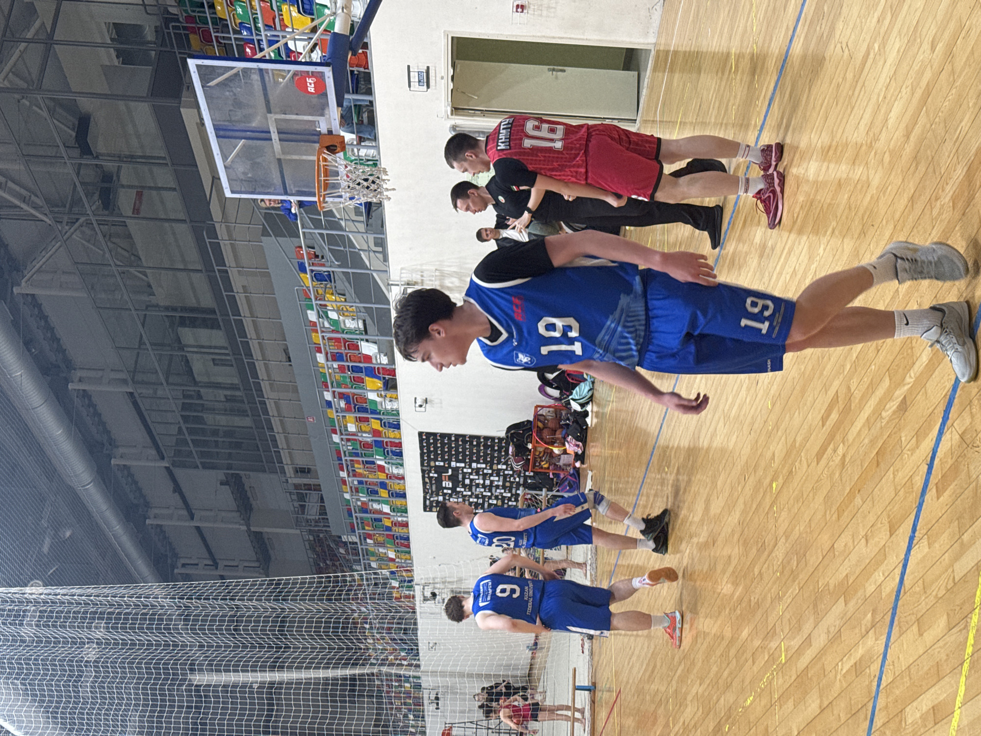 The image depicts a basketball court, likely indoors, with a view towards the stands and a basketball hoop. Several individuals are present, most of whom appear to be basketball players in uniforms. They wear blue and red jerseys, and numbers such as "19" and "16" are visible. Some individuals appear to be coaches or referees. The setting seems to be a sports facility or arena in Kazan, Russia, based on the jerseys.