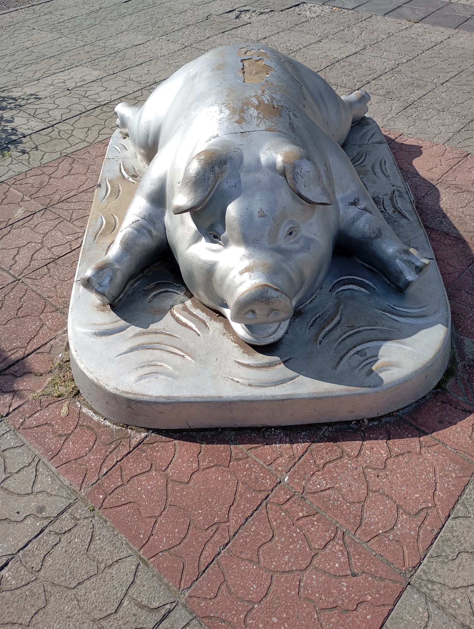 A silver sculpture of a pig lies on its back on a decorative concrete slab on a paved sidewalk in Horishni Plavni, Ukraine. The pig sculpture is detailed, with visible ears, snout, and legs. The concrete slab it rests on has wavy decorative lines. The sidewalk is composed of interlocking square tiles, some red and some gray, with a cracked pattern. Shadows cast by the sculpture and possibly nearby trees fall across the sidewalk. The scene appears to be outdoors in daylight.