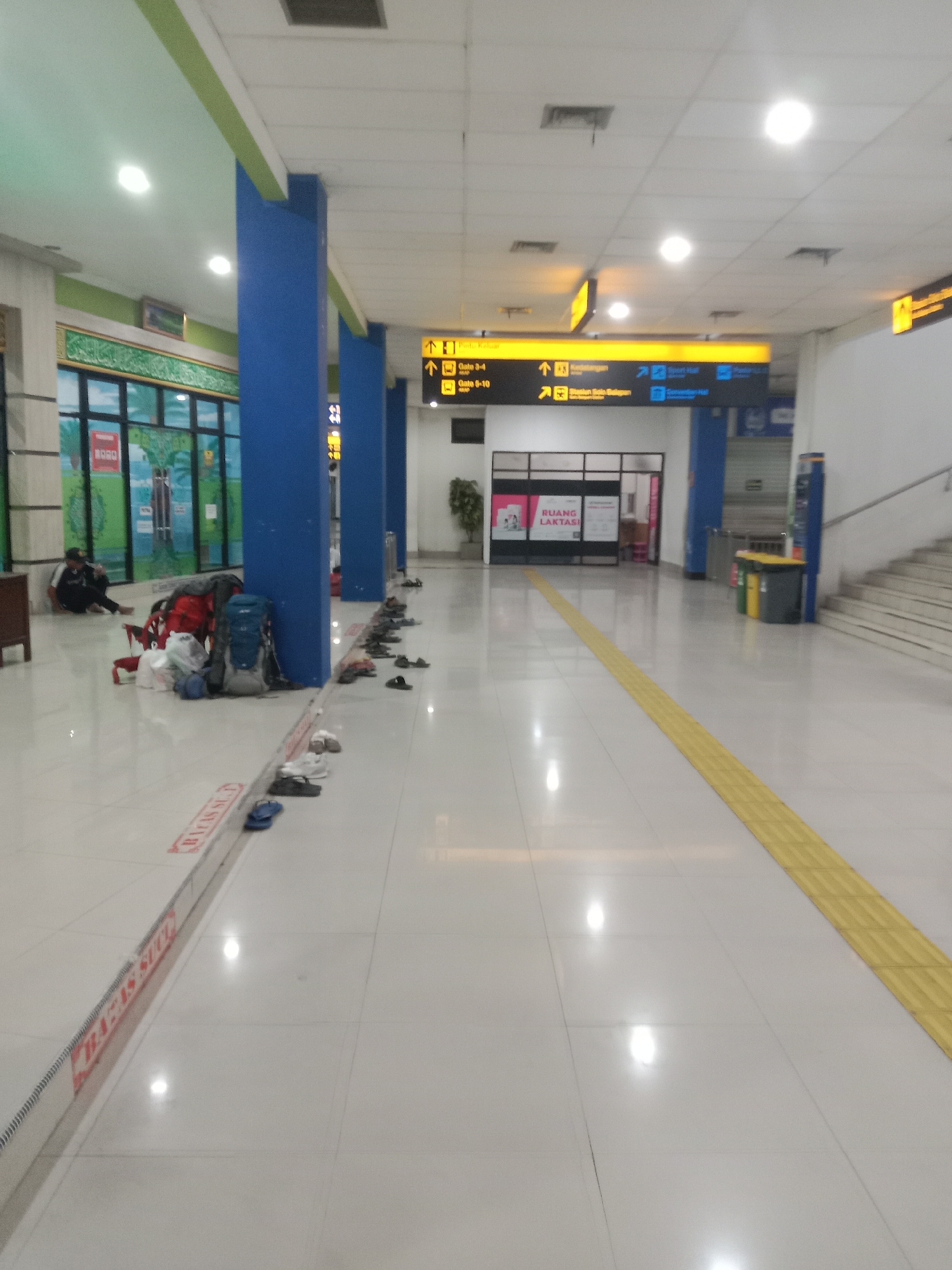 The image depicts a long, well-lit corridor within what appears to be a transportation hub, likely an airport or train station, in Surakarta, Indonesia. The floor is tiled in a light color, creating a reflective surface that bounces the light from numerous ceiling fixtures. Along the left side of the corridor, a person is seated on the floor, leaning against a wall, with several bags and backpacks near them. More luggage is piled on the floor further down this side, suggesting a waiting area or a space for travelers.
A prominent feature of the scene is the series of large blue columns that run vertically through the space, dividing the corridor. Above the columns, overhead signage with directional arrows and text in Indonesian ("Gate 3-4," "Gate 9-10," "Stasiun Solo Balapan," "Convention Hall") is clearly visible, indicating various destinations or facilities within the building. A yellow tactile paving strip runs along the right side of the corridor, a common safety feature for visually impaired individuals.
On the right side of the corridor, there are stairs leading upwards and a trash receptacle containing yellow and green bins. The walls are mostly white, with some areas featuring decorative panels or windows. The overall impression is one of a functional and somewhat sterile public space, populated by travelers and their belongings.