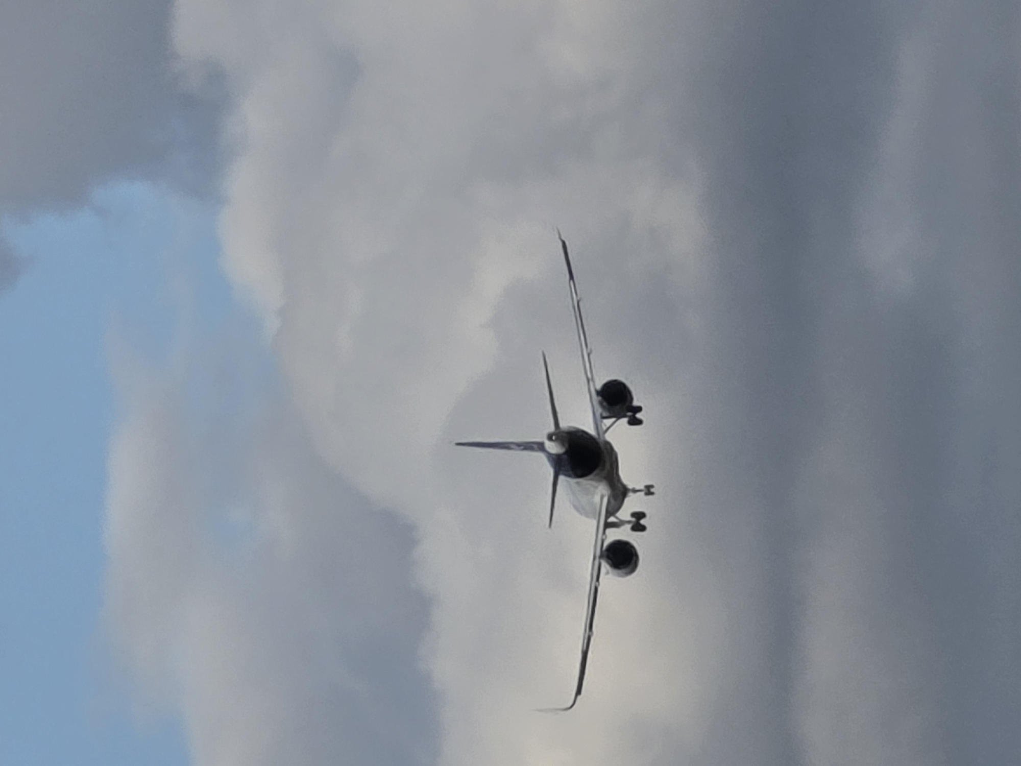 An airplane, viewed from the rear, is descending through a cloudy sky. The airplane is white with dark blue markings on its tail and engines. The landing gear is extended, indicating it is preparing to land. The sky is a mix of light and dark gray clouds with patches of blue visible. The image was taken in Flörsheim, Germany.