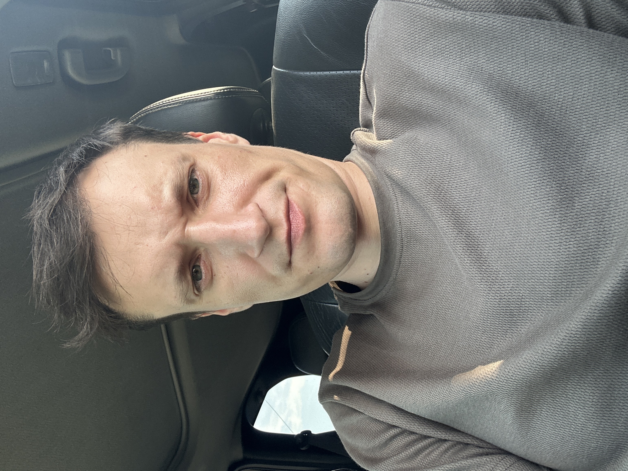 A medium shot shows a man sitting in a car. He is looking directly at the camera with a neutral expression. The man has short, dark hair and is wearing a grey long-sleeved shirt. The interior of the car is visible, including the dark leather seats and a sunroof. The background outside the car window shows a cloudy sky. The image appears to have been taken during the day.