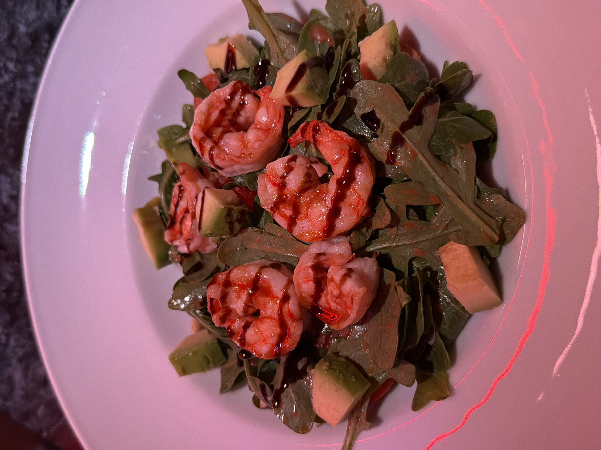 The image shows a close-up of a plate of food. The plate is white and contains a salad composed of arugula, avocado, and shrimp. A dark balsamic glaze is drizzled over the salad ingredients. The lighting in the scene appears to be artificial, casting a pink hue over the entire composition. The photograph was most likely taken indoors. It is impossible to know if the photograph was taken in the city of Chaikovsky, Russia.