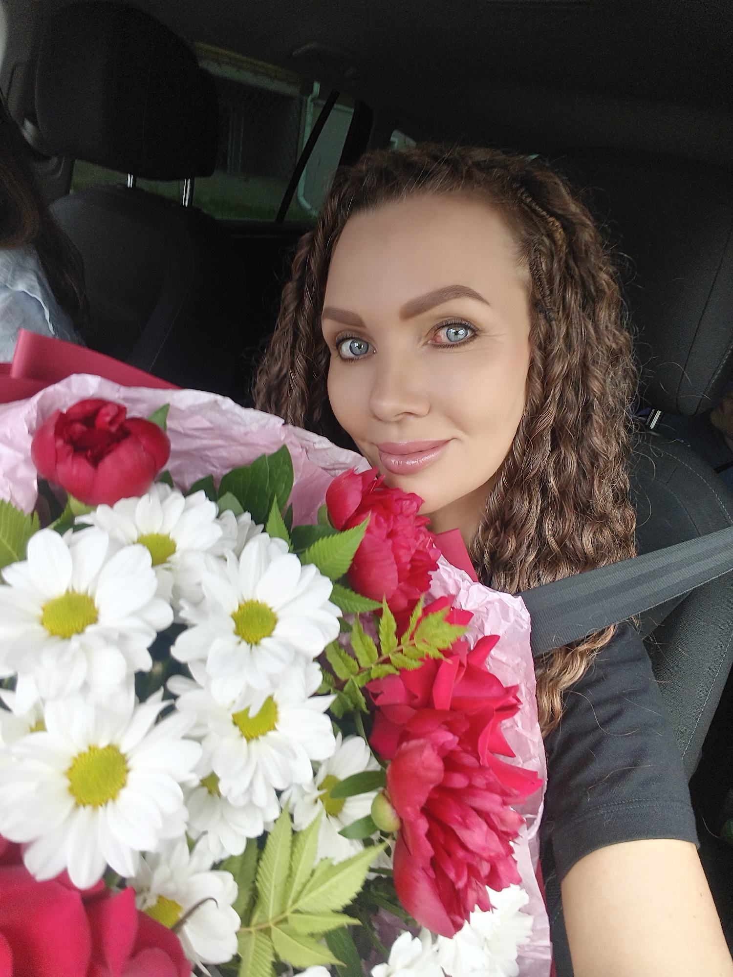 A woman with curly brown hair and blue eyes smiles at the camera, holding a bouquet of white daisies and red peonies in a car. She is wearing a black t-shirt and her seatbelt is fastened. The background shows the interior of the car and a glimpse of the outside.