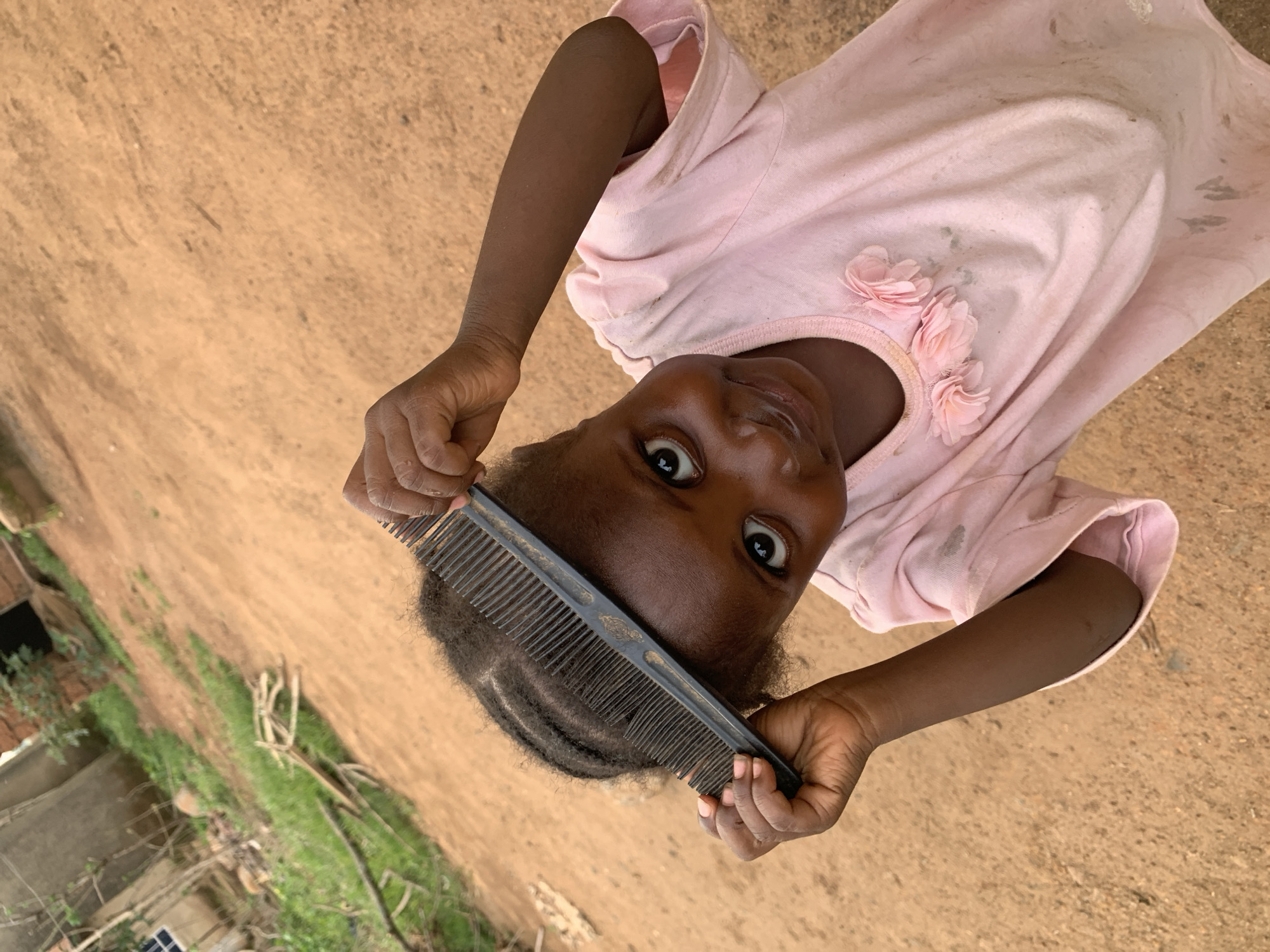 A young, dark-skinned girl, wearing a light pink shirt with flower embellishments, holds a black comb above her head, looking directly at the camera. She stands on a dusty, sandy ground. To the left of the frame is vegetation and a building. This image was taken in Agban, Nigeria.