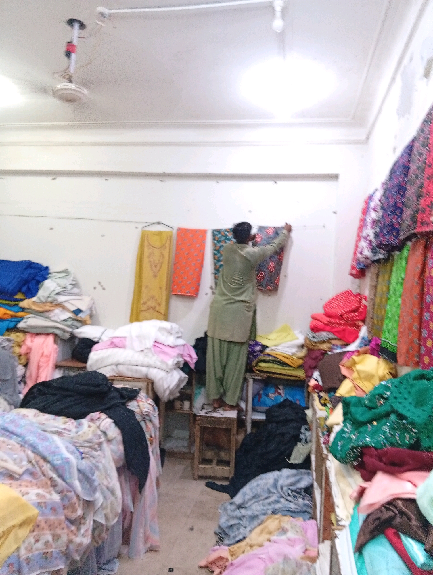 The image depicts the interior of a shop in Larkana, Pakistan, filled with textiles. A man, wearing a light green shalwar kameez, stands on a wooden stool, hanging a patterned fabric on a clothesline affixed to the wall. The shop appears to be a textile or clothing store, with piles of fabric in various colors and patterns stacked throughout the space. Bolts of fabric are visible on shelves and on the floor. Colorful garments hang from a rack on the right side of the image. The walls are white, and a ceiling fan is mounted overhead. The scene suggests a working environment where fabrics are stored, displayed, and possibly prepared for sale or tailoring.