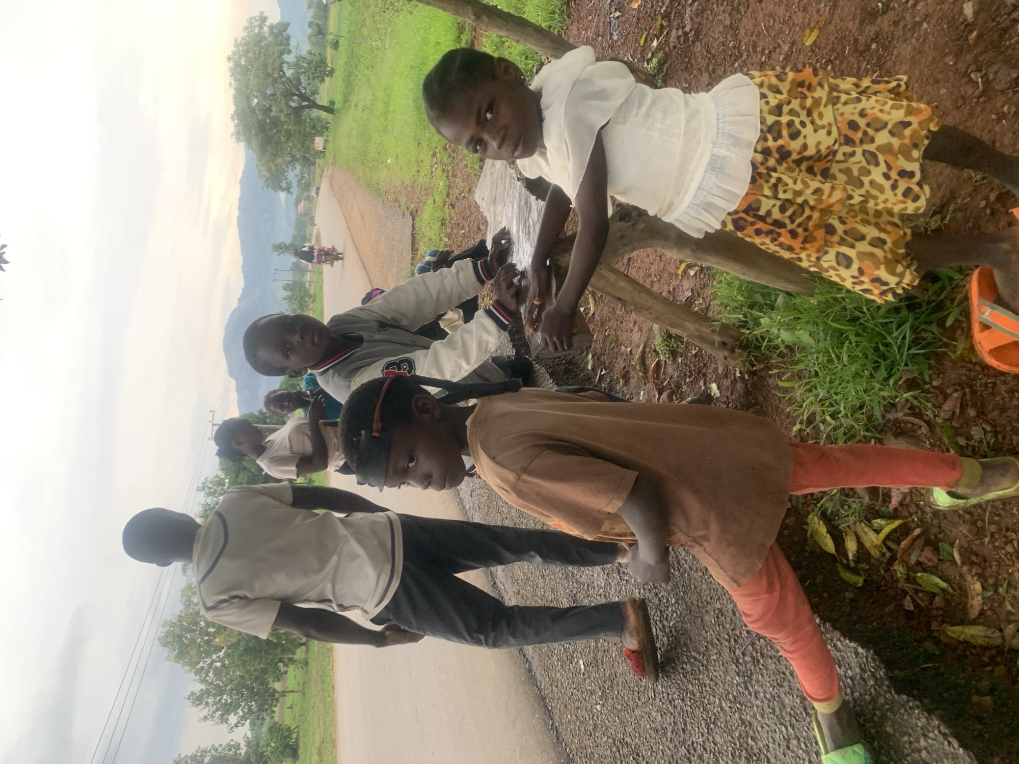 The image depicts a group of children and adults gathered outdoors in what appears to be a rural area. A young boy in a light-colored shirt and dark pants stands facing away from the camera, while several children are gathered around a rough wooden structure. One girl in a brown shirt and orange leggings is in the foreground, looking to the side. Another young girl in a white top and a patterned skirt is seated. In the background, a road can be seen with a motorcycle and a car in the distance. The landscape is characterized by green grass, trees, and hills in the background. The sky is overcast, suggesting a cloudy day. The overall scene suggests a casual gathering or interaction among the individuals.