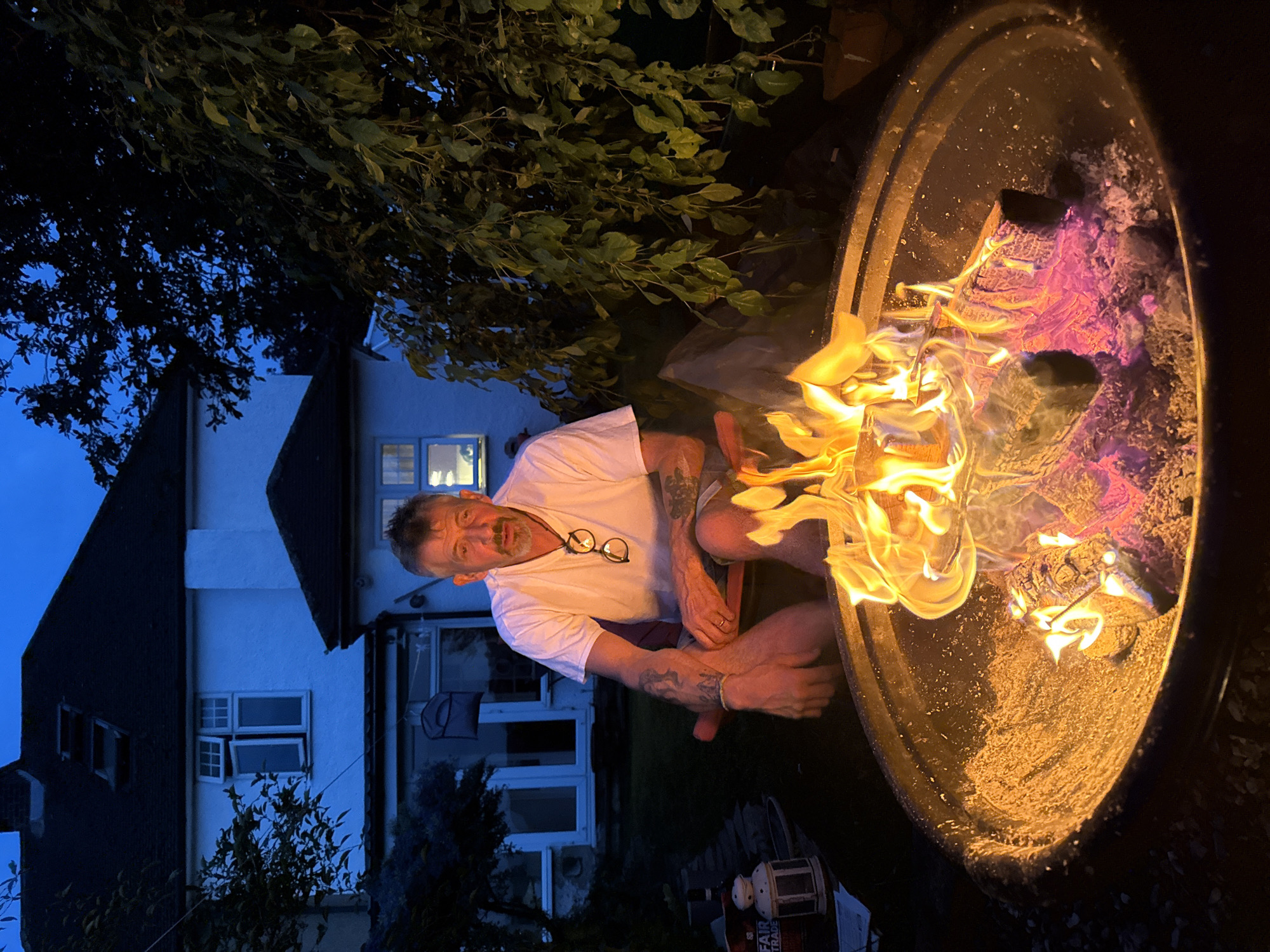 A man sits outdoors near a fire pit. He wears a white t-shirt and has glasses hanging from the neckline. He is sitting on a reddish-brown chair. The fire pit contains burning wood, which is the source of a bright yellow and orange light. Some logs are glowing with pink embers. Behind the man is a light-colored two-story house with a dark roof. A tree with green leaves is partially visible above the house. The sky is a deep blue, suggesting it is evening or night. A Fairtrade branded lantern is visible next to the fire pit. The location is identified as Stotfold, United Kingdom.