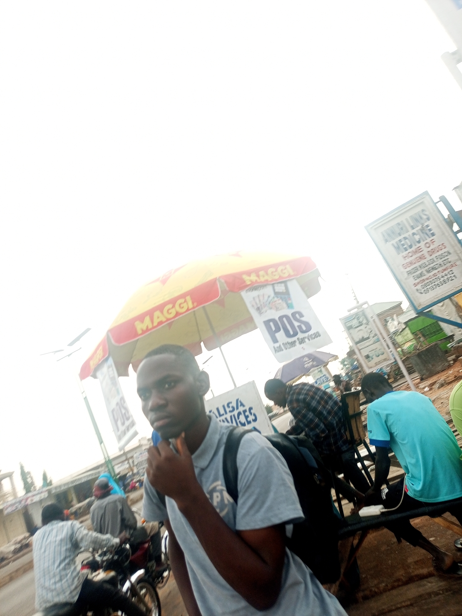 The scene depicts a street in what appears to be Tempure, Nigeria. A young man wearing a grey t-shirt and a backpack is in the foreground, holding something to his mouth. Behind him is a "Maggi" branded umbrella shielding a vendor selling "POS and other services." A sign reads "ANMURI LINKS MEDICINE HOME OF GENUINE DRUGS," including contact information. People are present, including some on motorbikes. The sky is bright, suggesting daylight.