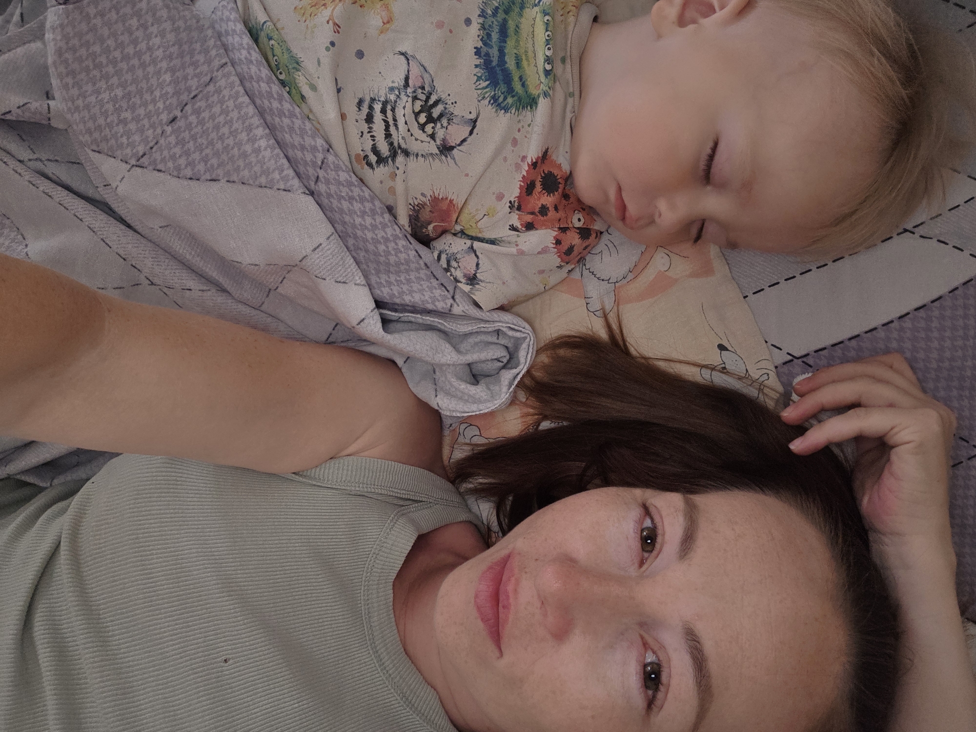 A woman and a sleeping child are lying in bed together. The child is on the left, with their eyes closed and head resting on a pillow. They are wearing a t-shirt with a colorful monster print and are partially covered by a gray and white patterned blanket. The woman is on the right, facing the camera. She has brown hair and is wearing a light green ribbed tank top. Her left hand is resting on her forehead. The background consists of bedding with a purple and white checkered pattern and a pillow with cartoon characters. The overall scene appears to be a domestic, indoor setting, likely a bedroom. The image does not provide any specific details or visual cues that would allow for the determination of its location within the city of Chaikovsky, Russia.