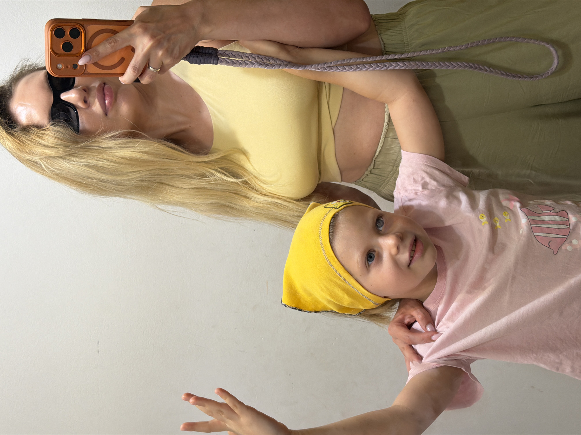 This is a medium shot, eye-level selfie taken indoors against a plain, light-colored wall. A woman with long blonde hair and sunglasses is holding up a phone in her right hand, taking the picture. She is wearing a yellow crop top and olive green pants. A young child, wearing a pink t-shirt and a yellow headscarf, stands in front of the woman, with their left arm raised. The woman's right hand is resting on the child's shoulder. The child is looking slightly to their right. The wall behind them has a faint horizontal marking. The setting appears to be an indoor changing room or dressing area within a shop, possibly a clothing store located in Budva, Montenegro, given the context.