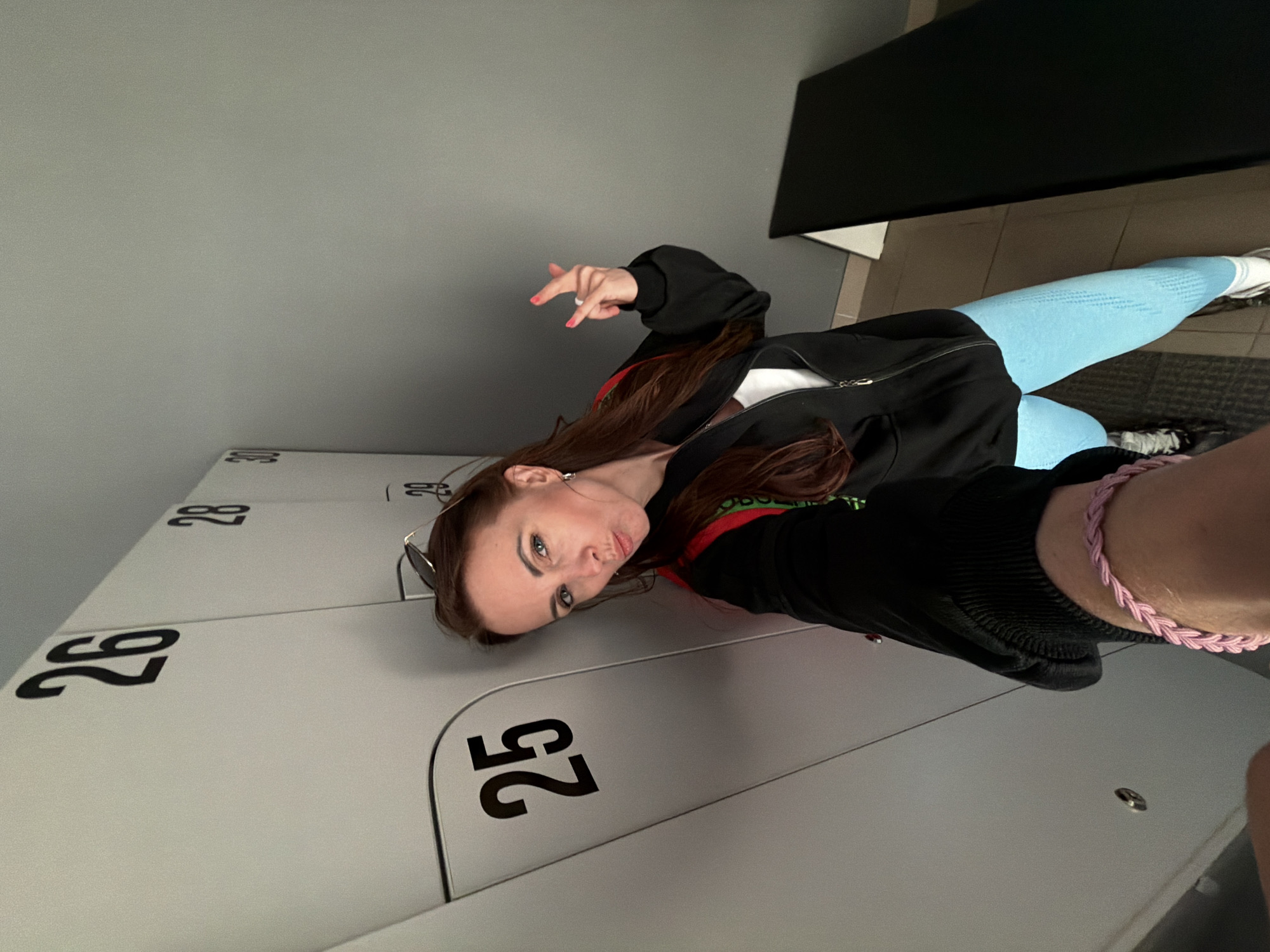 A young woman is captured in a locker room setting, likely in Birobidzhan, Russia. She has long, dark brown hair and is dressed in a black zipped hoodie over a white top, light blue athletic leggings, and white sneakers. Sunglasses are resting on her head. She is leaning against a row of light grey lockers, which are numbered "25," "26," "28," and "30" in black lettering, some appearing inverted due to the photo's perspective.
The woman is posing playfully for the camera, pouting her lips and making a "peace sign" or "rock on" gesture with her right hand, showcasing pink nail polish. A pink braided bracelet adorns her left wrist. The background includes a dark, rectangular object, possibly a bench or shelf, and a dark mat on the floor. The scene suggests an indoor environment with artificial lighting. The overall mood is casual.