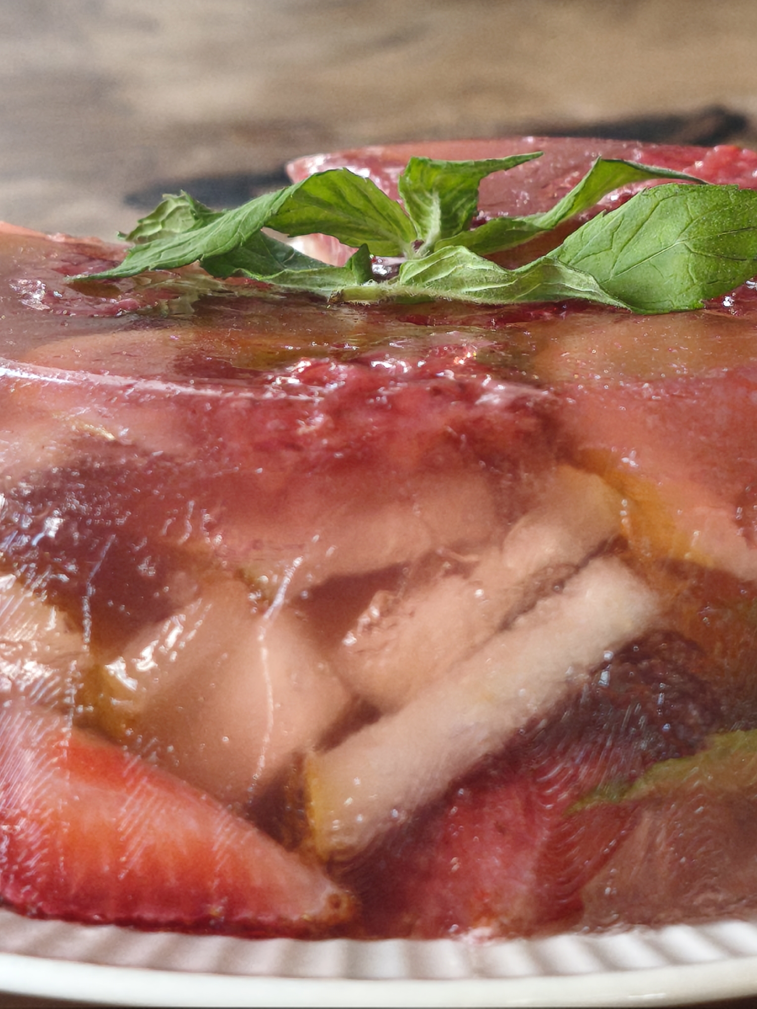 A close-up, overhead shot features a gelatin dessert filled with sliced strawberries and mint leaves. The dessert is presented on a white plate with a ridged edge. The background is a blurred wooden surface.