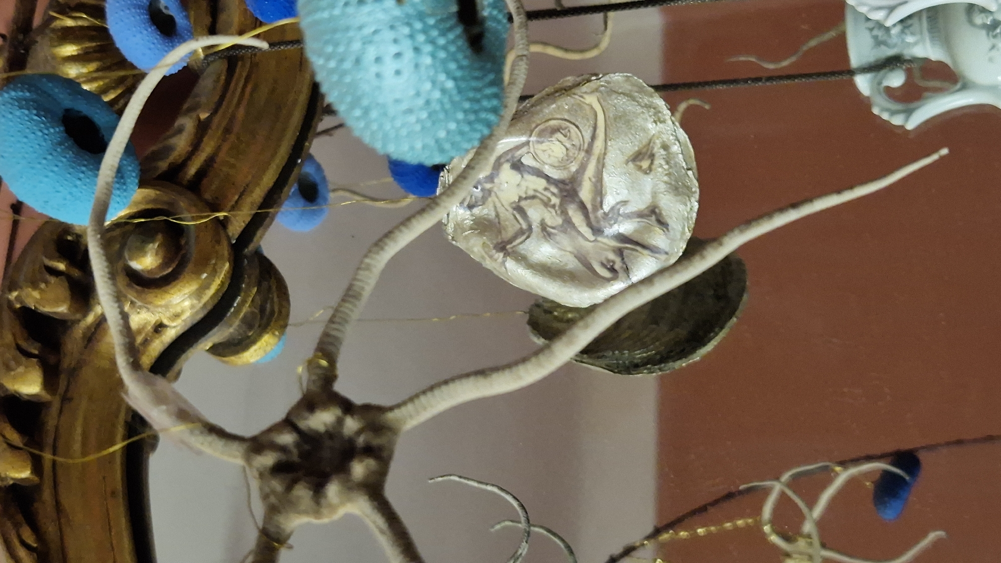 The image shows a close-up of an ornate, possibly antique, golden frame with decorative elements. Hanging from the frame are several objects: a starfish-like structure with long, thin arms, a dark shell, and a lighter, shell-shaped object with a detailed etching or painting of a human figure in motion. Several bright blue, textured, spherical objects are also attached to or hanging near the frame, possibly resembling sea urchins or decorative baubles. The background is a blurred reddish-brown surface. The overall impression is of a decorative display, possibly an art installation or part of a larger decorative piece. There is no indication of the city of Taranto, Italy, or any specific location within it in the image.