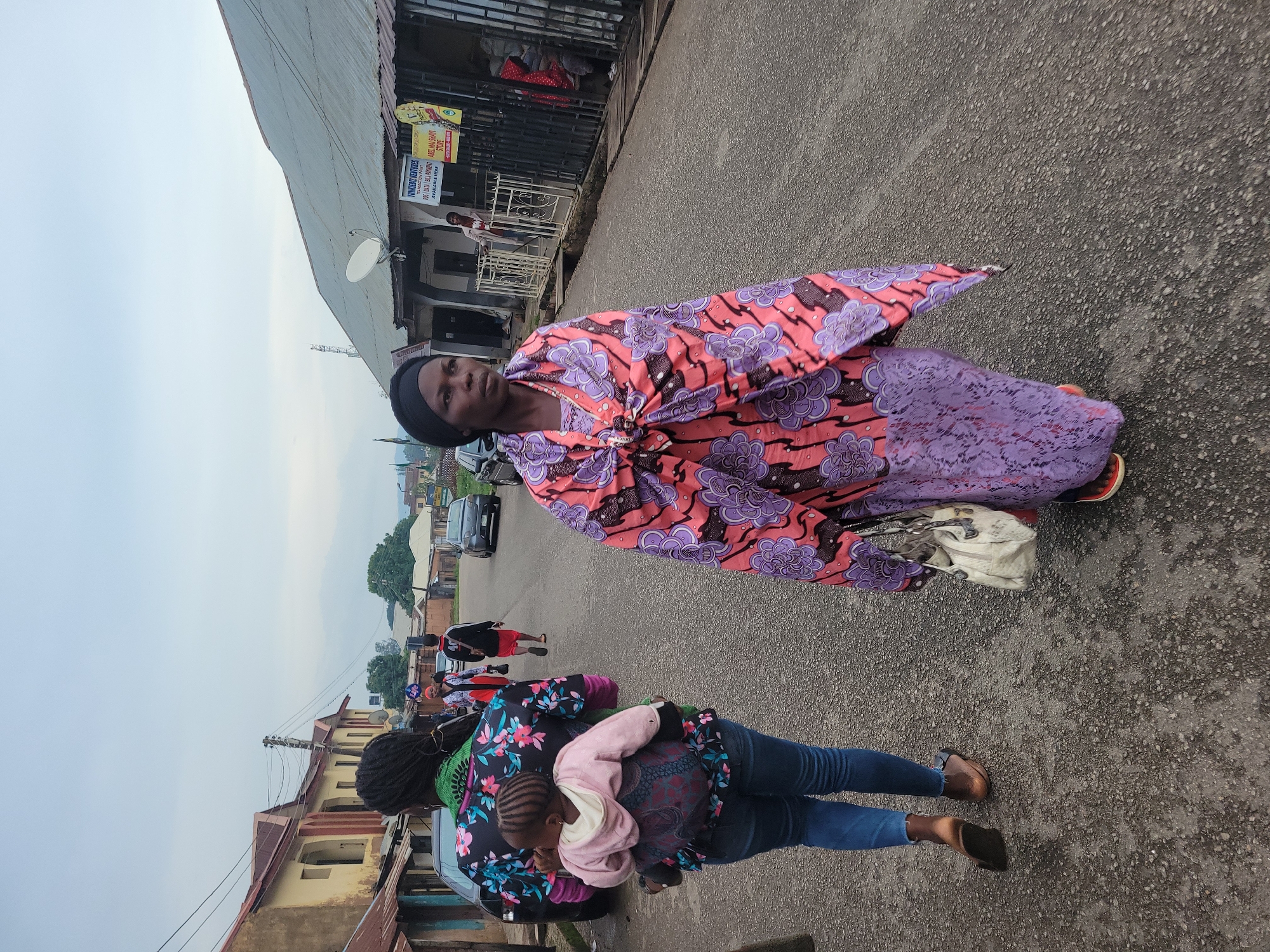 In Jos, Nigeria, on an overcast day, a woman walks down a paved street, her body covered in a vibrant pink and purple patterned fabric wrapped around her like a shawl and skirt. She wears a black headscarf and carries a textured white handbag. To her left, another woman walks away from the camera, also carrying a child on her back in a colorful fabric wrap. This second woman wears a brightly patterned top with flowers and a green collar, and blue jeans. A young child, wearing a pink hooded garment, is secured to her back. Several cars are parked or driving on the street. Buildings line the sides of the street, some with corrugated metal roofs and others with red-tiled roofs. The sky is a muted gray, suggesting the early morning or late afternoon. In the background, a sign is visible on one of the buildings. The overall scene depicts a typical street life in a Nigerian urban environment.