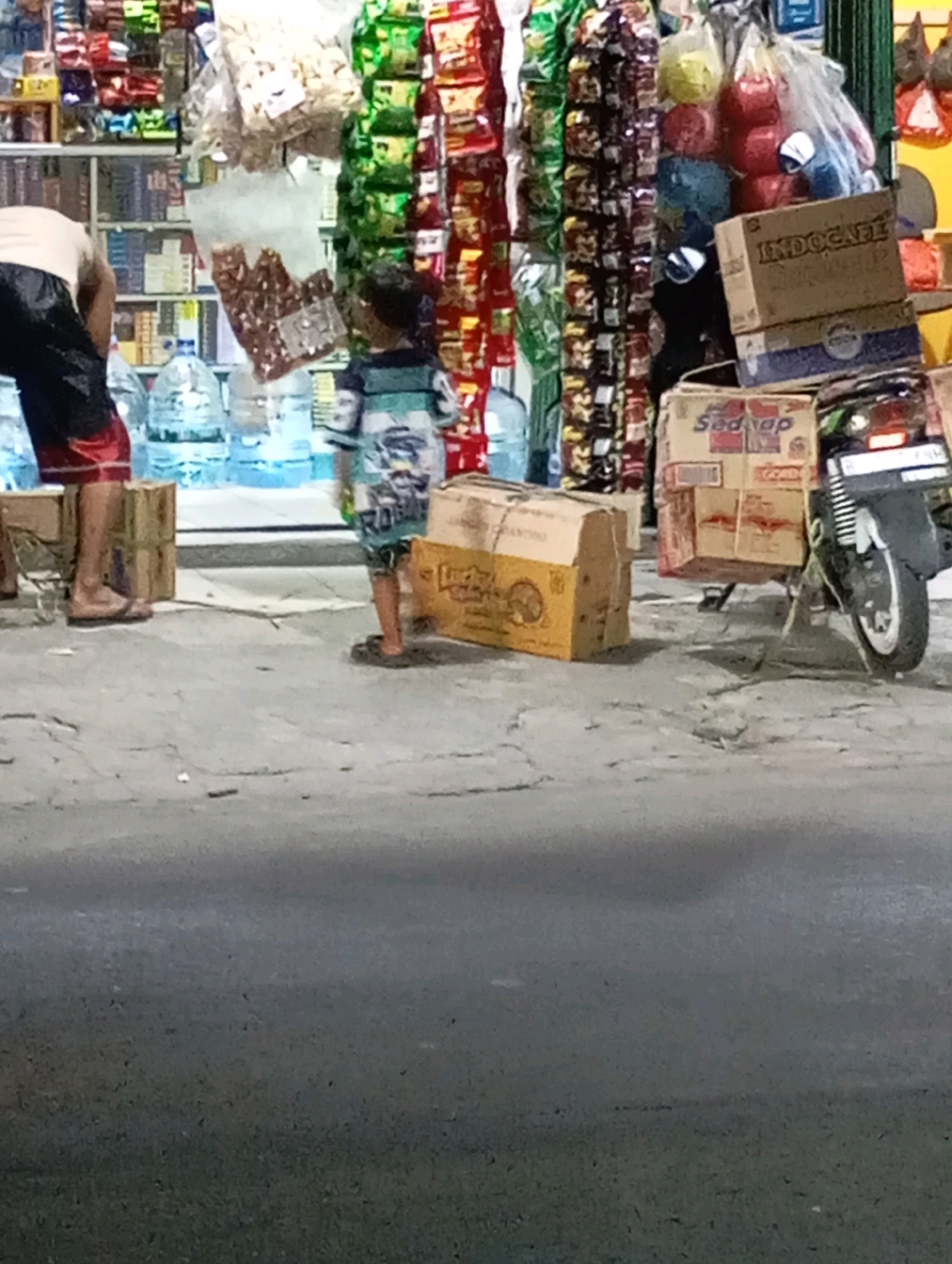 This image captures a street-level view of a store or market stall in Babelan, Indonesia. In the foreground, a person wearing a dark shirt and red and black patterned shorts stands with their back to the camera, facing a display of goods. Beside them, a child in a horizontally striped shirt and shorts is also facing away from the camera. Behind them, shelves are stocked with various packaged food items and drinks, including bottles of water. Several cardboard boxes are visible on the ground and stacked, some with visible branding like "INDOCAFE" and "Sedap." A motorcycle is parked to the right, with boxes secured to its rear. The scene appears to be outdoors, with a paved surface in the foreground and the stalls extending into the background.