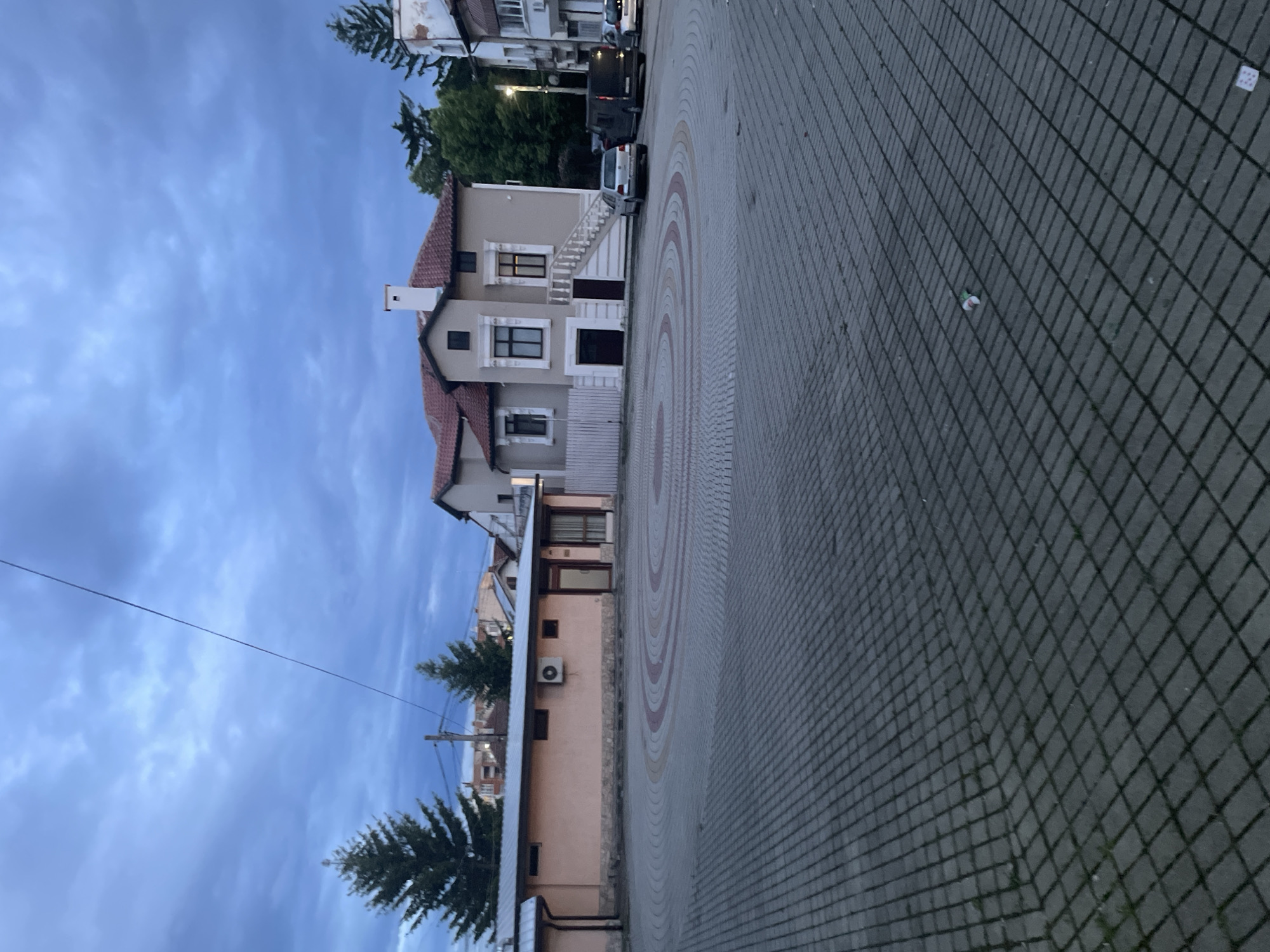 This image depicts a quiet street scene in Ihtiman, Bulgaria, captured during what appears to be late afternoon or early evening, given the soft, diffuse light and a sky filled with scattered, light-blue clouds.
In the midground, a prominent two-story house with light beige walls and a red-tiled roof stands out. It features a distinctive white chimney on the roof and multiple dark-framed windows. A white outdoor staircase with a balustrade leads up to an entrance on the upper floor. Behind this house, several parked vehicles are visible on what appears to be a residential street.
To the left of the main house, in the foreground, is a lower, single-story building with a flat roof and an air conditioning unit mounted on its peach-colored wall. Further left, tall evergreen trees, likely pine or fir, add greenery to the scene, and a utility pole with a power line is also visible against the sky.
The ground in the foreground and midground is paved with interlocking tiles. The area closest to the buildings features an elaborate circular pattern in shades of grey, red, and beige, while the broader right side of the paving consists of a uniform grid of grey square pavers. A small piece of litter, possibly a discarded cup, can be seen on the grey paved section.
No human activity is explicitly depicted, lending a tranquil atmosphere to the scene. There is no visible text or signage in the image.