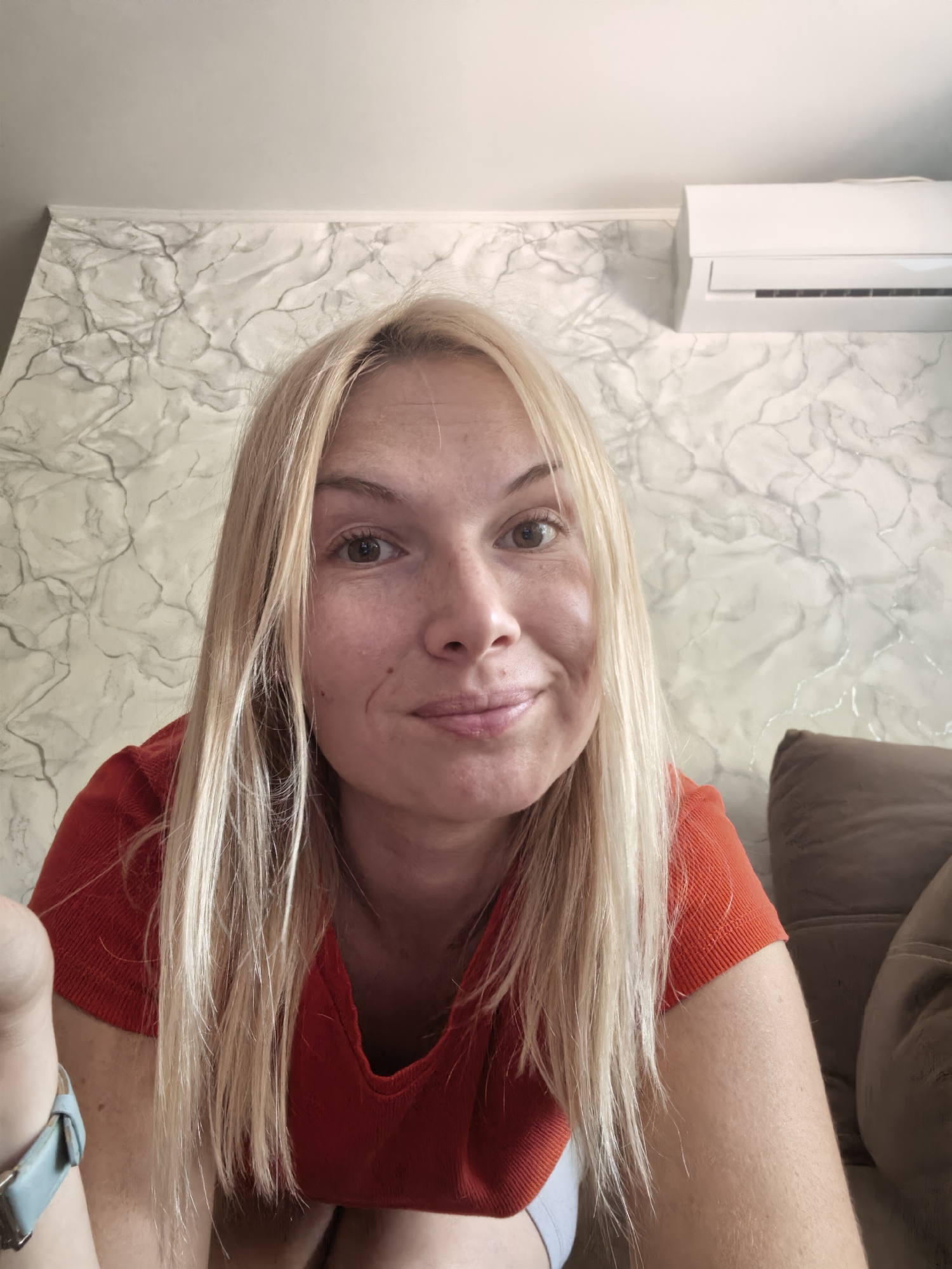 A blonde woman in a red shirt is pictured from the chest up, looking directly at the camera with a slight smile. She has fair skin with some freckles and light brown eyes. Her long, blonde hair is worn down, framing her face. Behind her, a textured, light-colored wall with grey veining is visible, along with a white air conditioning unit mounted high on the wall. To the right, a portion of a brown upholstered couch is present. On the left side of the image, a hand with a light blue watch strap is partially visible. The scene appears to be indoors.