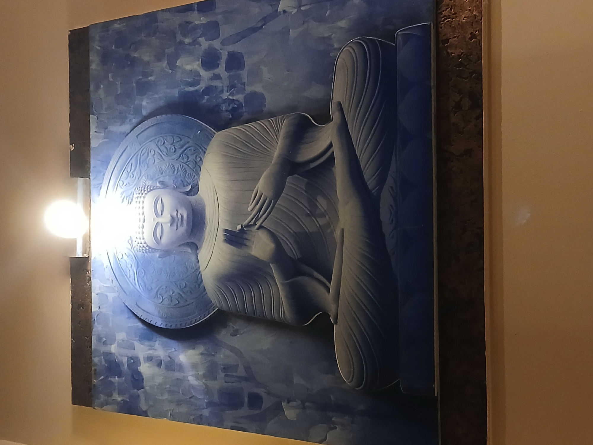 A framed artwork of a reclining Buddha is mounted on a wall, possibly in an indoor setting in Bengaluru, India. The Buddha figure is depicted in a reclined position with its hand resting on its thigh, its head surrounded by a large halo. The artwork has a blue and grey palette with a mottled background reminiscent of brushstrokes. The frame is a dark color, with a textured finish visible on the right side. A light source positioned to the left illuminates the artwork, creating glare on the surface. The wall behind the artwork is painted in a light beige color.