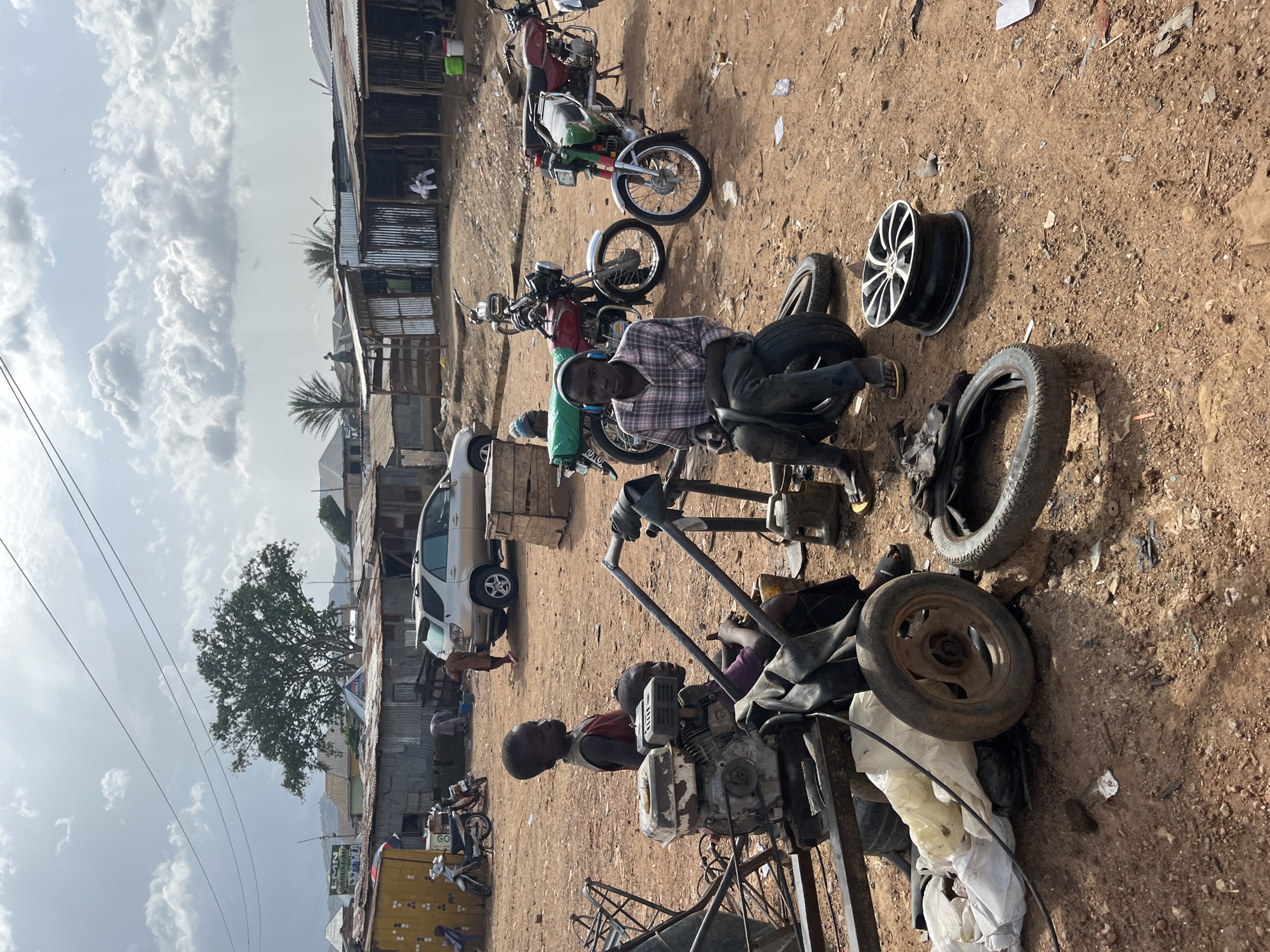 This is a medium shot of a busy outdoor scene, likely in the city of Jos, Nigeria. In the foreground, a young man wearing a plaid shirt and headphones sits on a tire, holding another tire. To his left, two younger children are near a stationary engine. Scattered around them are various tires, a car rim, and a large white bag. In the mid-ground, several motorcycles are parked, including a red one and a green and red one. Behind the motorcycles, a silver SUV is parked. The background shows a collection of corrugated metal structures, typical of market stalls or informal housing in Jos. The ground is dusty red earth, and overhead, a cloudy sky suggests a warm climate. The scene conveys a sense of activity and daily life in a developing urban area.