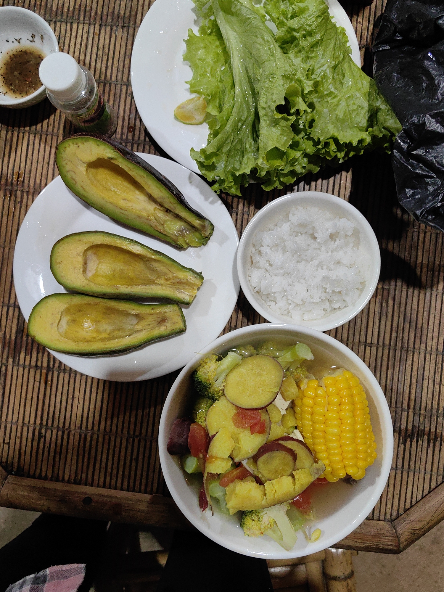 A still-life photograph depicts a meal arranged on a bamboo placemat, possibly in Xã Đàm Thủy, Vietnam. The meal consists of three avocado halves on a white plate, a bowl of white rice, a bowl containing what appears to be vegetable soup with corn on the cob, sliced sweet potatoes, broccoli, and other vegetables, and a plate of lettuce leaves. A small bowl with a sauce and a spice shaker bottle are also visible. A black plastic bag partially obscures the upper right corner of the frame. The scene is lit from above.