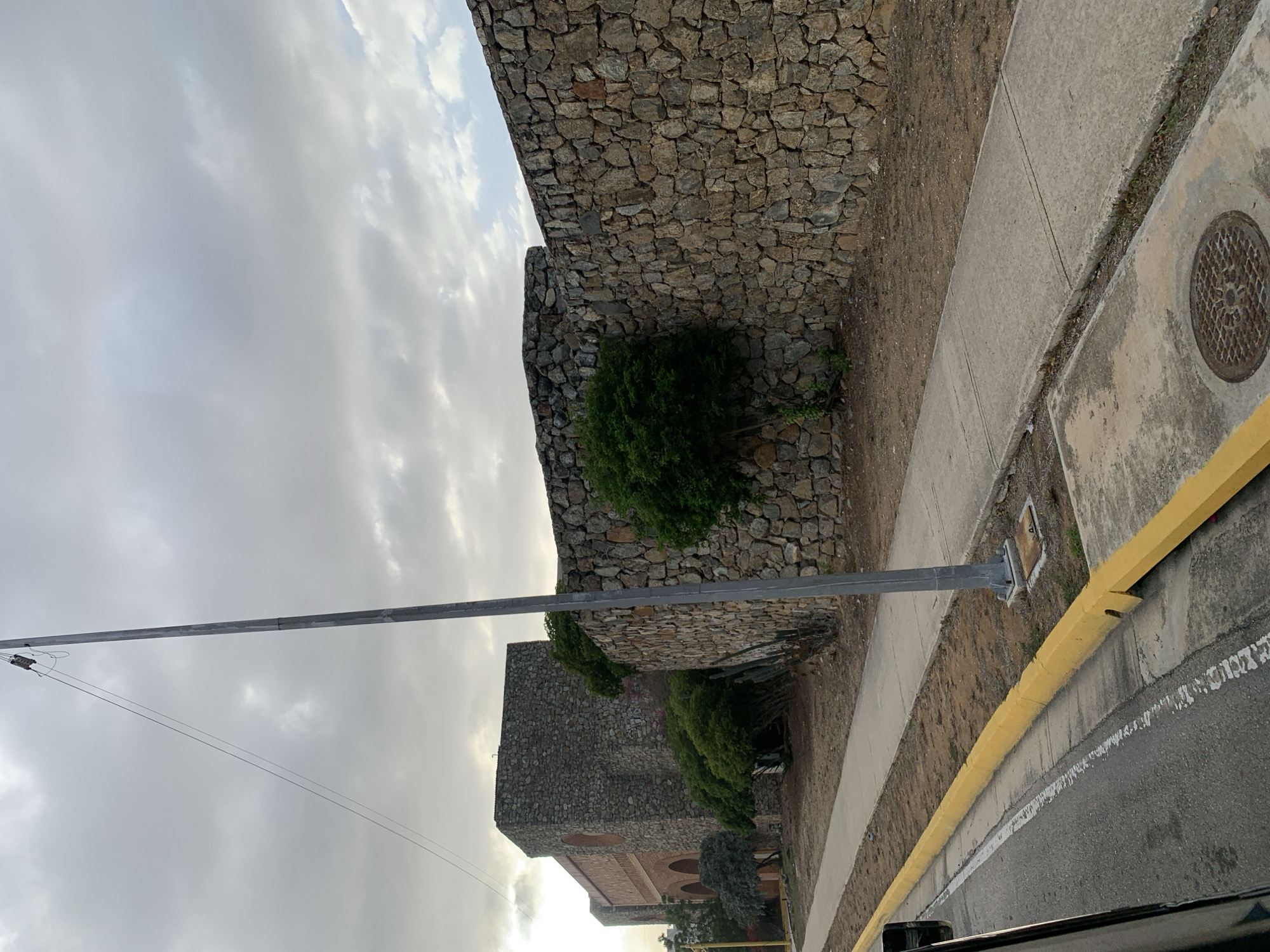 This media file presents an outdoor scene in Porlamar, Venezuela, showcasing a road running alongside a historic-looking stone wall under a cloudy sky. The photograph appears to be taken from inside a vehicle, with a glimpse of a dark car window frame in the bottom right corner.
The primary subjects are two prominent stone walls, built from irregular, rugged-looking gray and brown stones. The wall in the foreground is tall and sturdy, with several green bushes and sparse vegetation growing along its base and partially climbing its surface. Behind this, another stone structure, possibly a building, is visible, featuring arched openings that suggest historical architecture. More green foliage is present around the base of this second structure.
Parallel to the main stone wall, there is a strip of reddish-brown earth, followed by a concrete sidewalk. A circular metal manhole cover with a patterned surface is embedded in the sidewalk on the right side. Beyond the sidewalk lies an asphalt road, which includes a clear white line marking a lane and a distinctive long, bright yellow concrete curb or barrier. A tall, slender gray metal pole, likely a utility pole, stands vertically near the road, extending towards the sky, with thin wires running from its upper section.
The time of day appears to be late morning or afternoon, characterized by diffused light filtering through a predominantly overcast sky filled with soft gray and white clouds. The weather is cloudy and calm. There are no people visible, and no specific activity is taking place, rendering the scene quiet and still. No discernible text is present in the image. The stone construction and tropical-like vegetation serve as location cues consistent with the provided context of Porlamar.