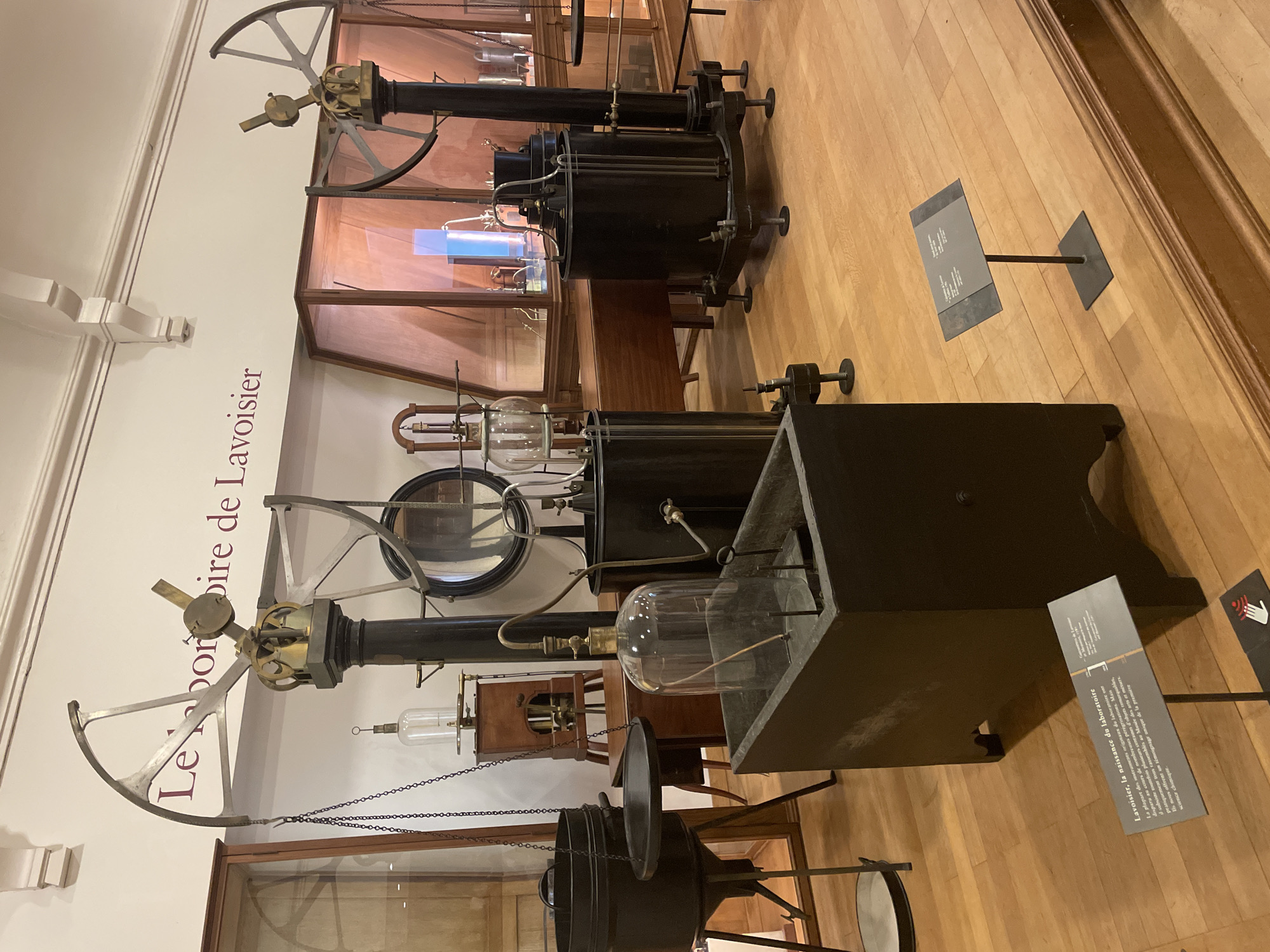 This image showcases a collection of historical scientific apparatus, likely displayed in a museum setting in Paris, France. The central focus is on two elaborate, dark-colored machines with prominent brass and metal components, including large, spoked wheels and glass vessels. These instruments appear to be part of an exhibition dedicated to the scientist Lavoisier, as suggested by the text on the wall which reads "Le laboratoire de Lavoisier" (Lavoisier's laboratory).
The scene is indoors, with a polished wooden floor. The lighting suggests a well-lit exhibition space, possibly with display cases containing other historical artifacts in the background, some of which are partially visible. One of the machines features a large cylindrical chamber, while the other has a more complex arrangement of tubes and chambers, including a prominent spherical glass container.
Several informational plaques are visible, including one in the foreground on the right that provides text in French. The text on the plaque translates to "Lavoisier, the birth of the laboratory," and it details the journeys of the original laboratory instruments and the preservation of hundreds of remarkable objects, offering a unique testament to the early days of chemical science.
The overall impression is one of historical preservation and scientific education, highlighting the foundational work of Lavoisier in the field of chemistry. There are no people visible in the image, and the scene is static, focused on the inanimate objects. The atmosphere is scholarly and informative.
