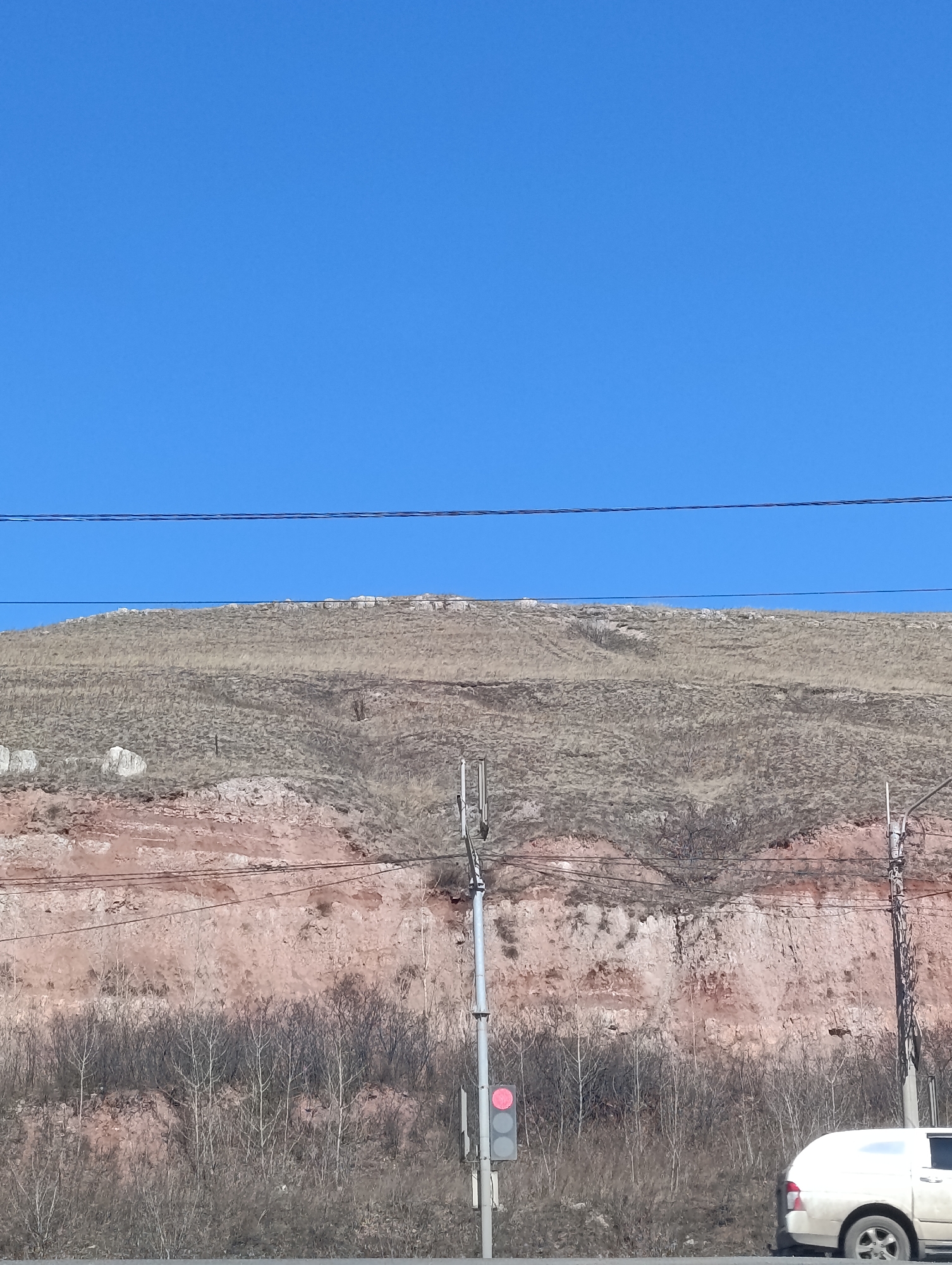 The image shows a view in Krasnoyarsk, Russia, of a hill under a clear blue sky. The hill exhibits two distinct layers: the top is covered with dry grasses, while the lower section reveals pinkish-red soil. A cluster of bare trees and shrubbery grows near the base of the hill. Power lines traverse the scene horizontally near the top of the image. A traffic light showing a red light and street poles are visible along the road in the foreground, and a white vehicle is parked beside the road.