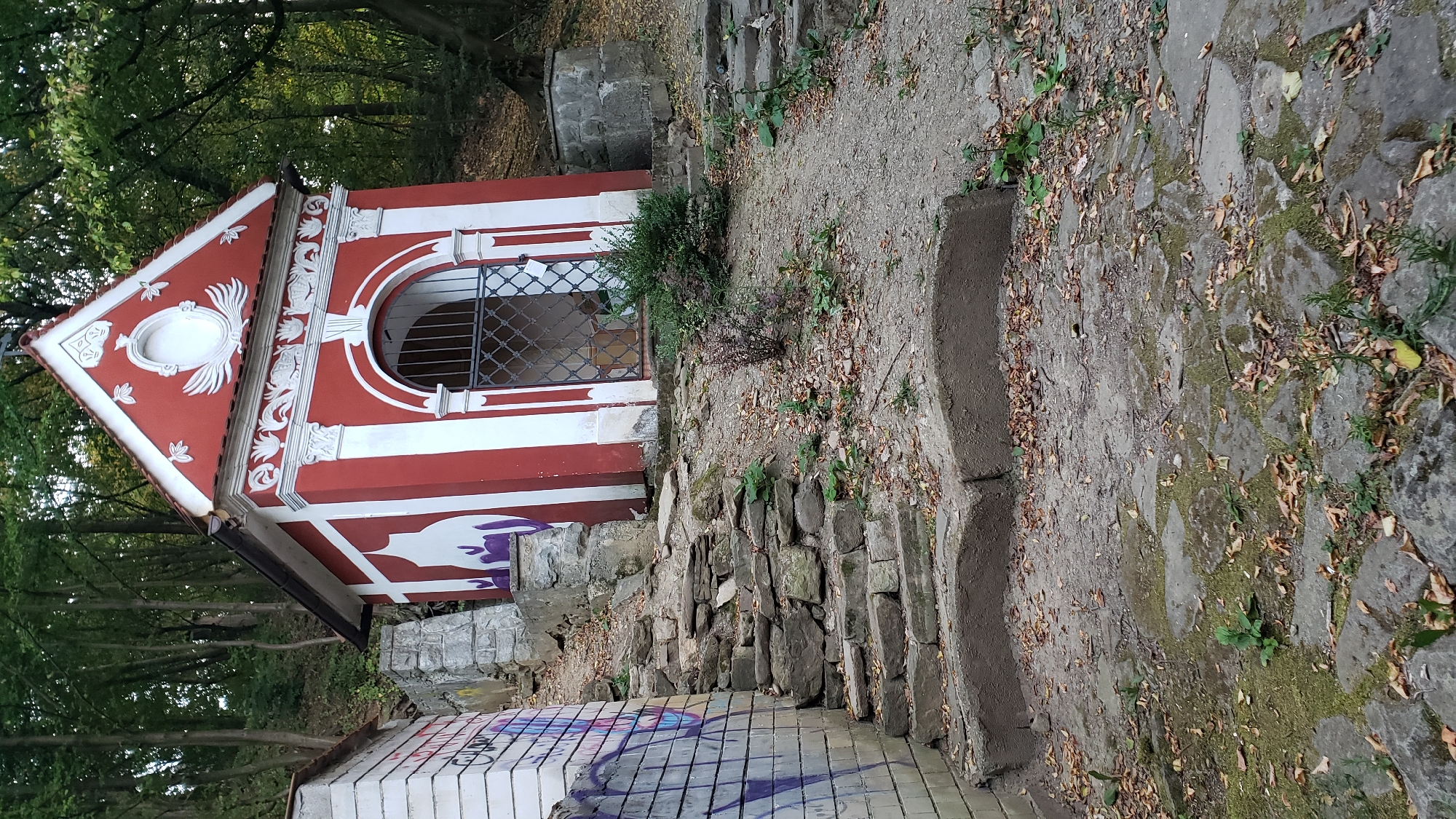 The image depicts a red and white chapel situated on a sloped, wooded terrain. The chapel features an arched entryway with a metal grate and decorative white trim. To the left of the chapel, a graffiti-covered white brick wall with a set of stone stairs leading upwards is visible. The ground is covered with dirt, scattered leaves, and some vegetation. The overall scene suggests a somewhat weathered and secluded location. This chapel is part of the Calvary in Prešov, Slovakia.