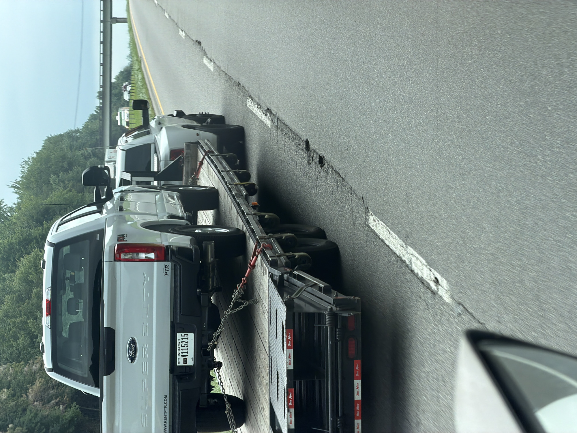 An outdoor, medium shot captures a light-colored Ford Super Duty truck being towed on a flatbed trailer on a highway. The truck's rear is visible, showing its license plate, which reads "INDIANA 4115215". The flatbed trailer is attached to another vehicle, partially visible in the foreground, and is secured by chains. Trees line the right side of the highway, and a bridge can be seen in the distance. The sky is overcast. The scene appears to be in Olin, United States.