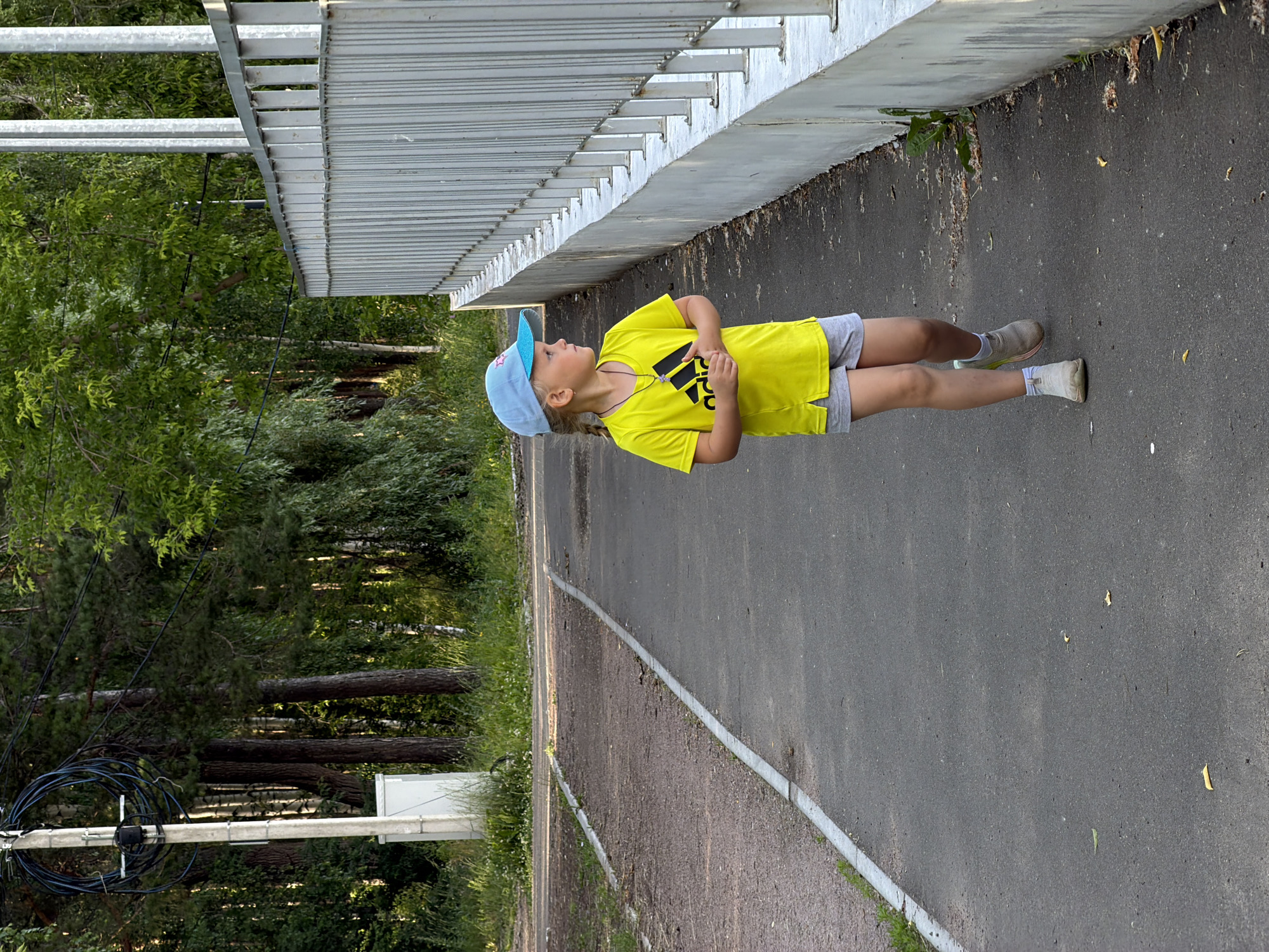 The image shows a young girl, wearing a yellow t-shirt, grey shorts, a blue cap, and grey sneakers, walking on a paved path next to a metal fence. She has blonde pigtails and is looking to her right. The path appears to be in a park or recreational area, with trees and foliage visible in the background. A concrete utility pole with coiled wires is also present on the left side of the image. The scene is illuminated by natural light, suggesting it is daytime. Based on the typical landscape and urban infrastructure, this scene could be located in the city of Chaikovsky, Russia, possibly in a park such as the one along the Kama River.