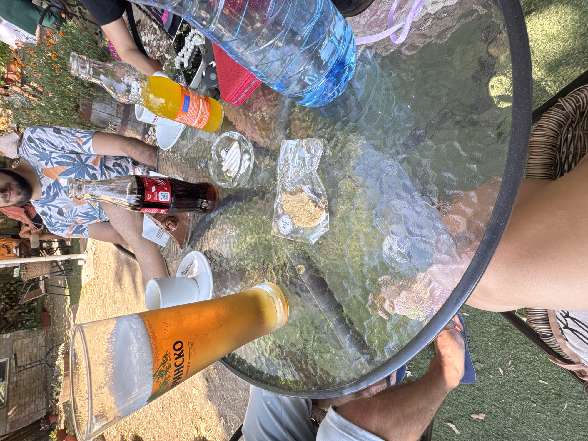 A group of people are seated around a round glass-top table outdoors, likely at a cafe or restaurant in Zheravna, Bulgaria, given the context of the prompt. The table is covered with various items including a tall glass of beer, a Coca-Cola bottle, a bottle of orange soda, a white coffee cup and saucer, an ashtray filled with cigarette butts, a small package containing a cookie, a red wallet with a zipper, and a large plastic bottle of water. The ground around the table is covered in grass and dry leaves, and in the background, there are chairs, tables, and foliage, suggesting an outdoor seating area. One person's legs and feet are visible in the foreground, wearing flip-flops, and another person's legs are also visible. A man with a beard, wearing a patterned shirt, is seated at the table. Other individuals are partially visible in the background. The lighting suggests it is daytime and sunny.