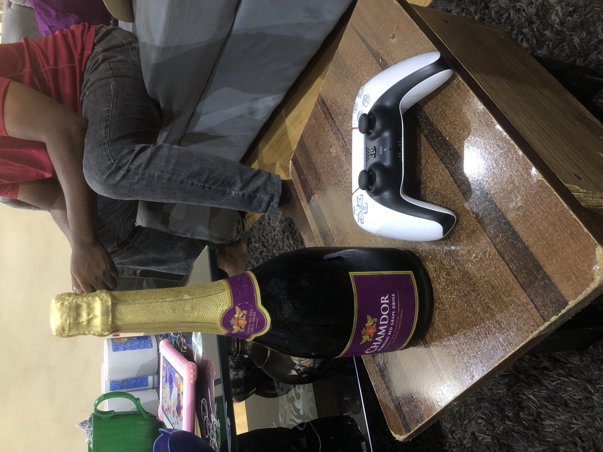 In a Lagos, Nigeria home, a bottle of Chamdor Sparkling Red Grape Drink, capped with gold foil, stands on a wooden table. Next to it lies a white PlayStation 5 controller. In the background, a person wearing a red shirt and grey jeans sits on a grey couch, holding a green cup. The scene suggests a relaxed evening of gaming and refreshments.