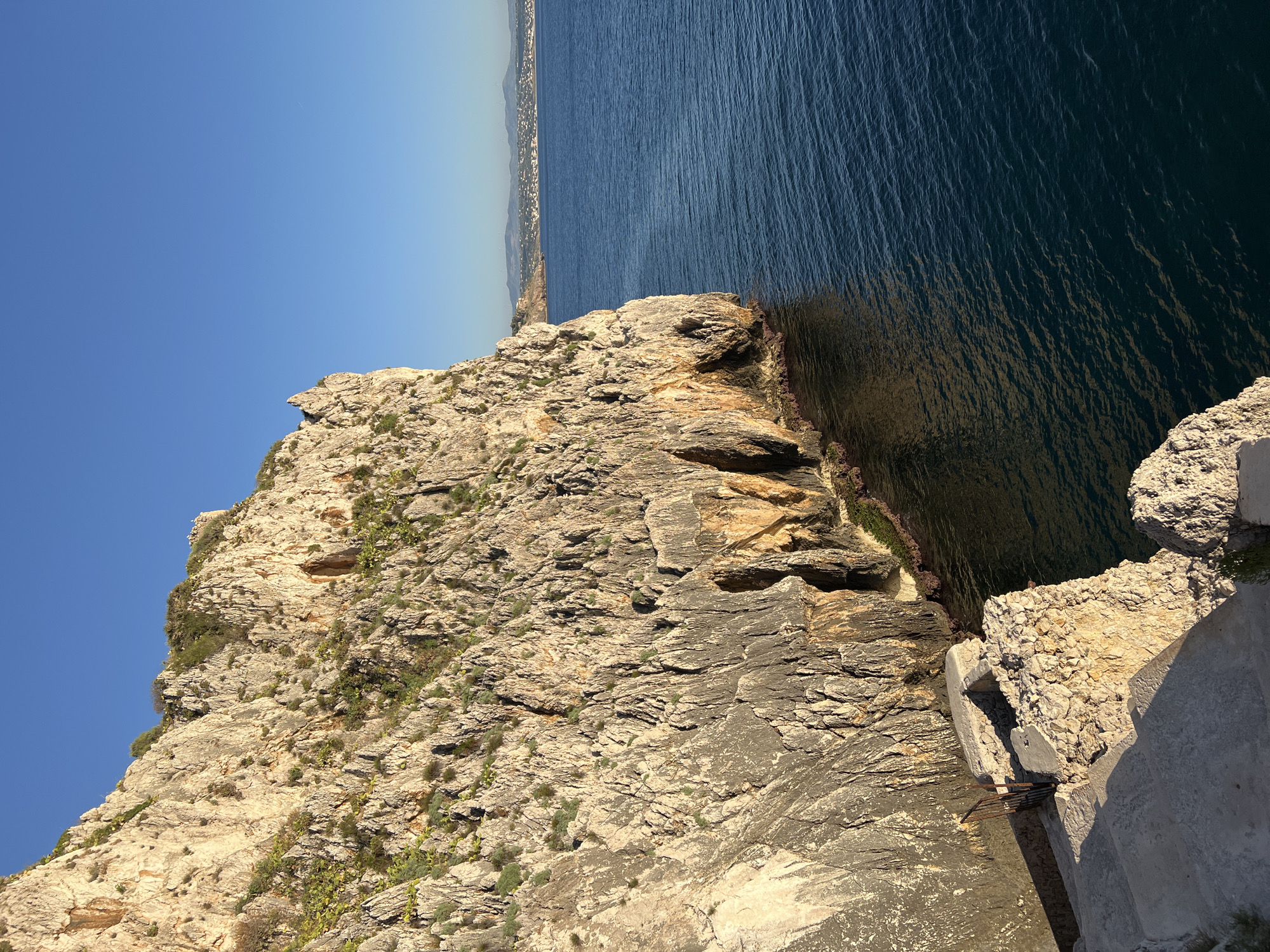 The image depicts a dramatic cliff face overlooking a deep blue sea, likely located in Terrasini, Italy, a coastal town known for its rugged beauty. The cliff, composed of layered rock, is textured with cracks, crevices, and patches of hardy green vegetation. Sunlight highlights the warm tones of the rock, casting shadows that accentuate its ruggedness.
In the foreground, a set of worn, concrete steps descends along the edge of the cliff. A small, weathered stone structure with a rusty metal gate is situated at the top of these steps, suggesting a pathway or an old entrance. The sea is calm, with gentle ripples reflecting the clear blue sky. In the distance, a hazy coastline is visible, dotted with buildings and possibly wind turbines, indicating the proximity of a populated area. The overall scene is one of natural grandeur and a sense of remote beauty, characteristic of the Sicilian coastline.