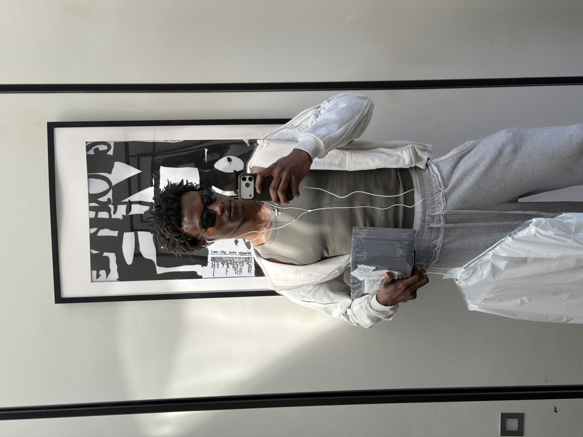 The image is a photograph taken in what appears to be an indoor setting, possibly a residential or commercial space in London, United Kingdom. The scene is captured in a mirror reflection. A Black man is the central subject, taking a selfie with his mobile phone. He is wearing sunglasses, a sleeveless shirt, a grey zip-up hoodie, and grey sweatpants. He has earphones plugged into his phone.
He is holding a stack of wrapped books or documents in one hand and a partially visible plastic bag in the other. Behind him hangs a framed black and white abstract art piece on a white wall. The top and bottom edges of the mirror are visible, indicating the reflective nature of the photo. The color palette is dominated by whites, greys, and blacks. The location can be inferred as being in London based on the UK delivery label on the package. A light switch or electrical outlet is visible near the bottom of the frame. The overall setting appears minimalist and modern.
