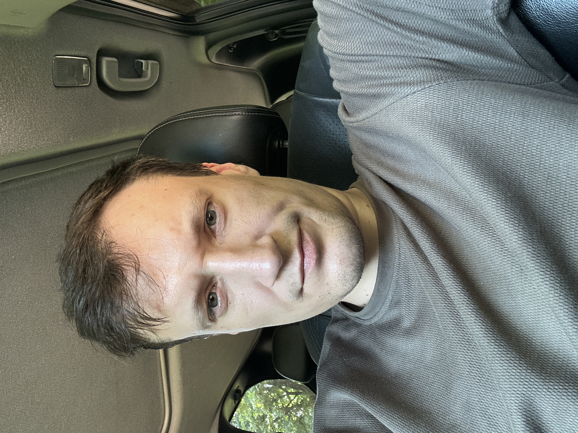 A medium shot photo shows a man with light skin, dark hair, and blue eyes sitting in the driver's seat of a car. He is wearing a grey t-shirt. The background includes the interior of the car with a sunroof and black leather seats, as well as trees visible through the rear window. The man is looking directly at the camera with a neutral expression. The lighting suggests it is daytime, possibly outdoors in a wooded area. This image does not show the city of Shatava, Ukraine.