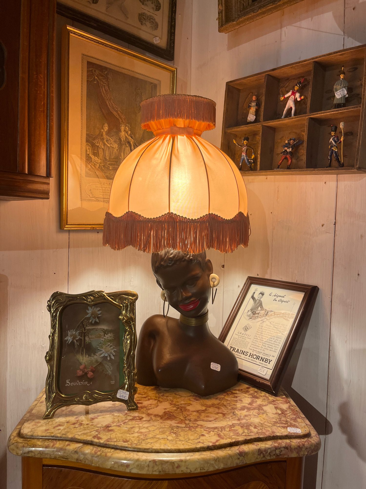 The image shows a display in what appears to be an antique shop or curio store in Yvoire, France. In the foreground, on a marble-topped table, is a decorative lamp with an orange, fringed shade. Beneath the lamp is a bust of a dark-skinned woman with red lips, large earrings, and a gold necklace. To the left of the bust is a small, ornate gold picture frame containing a painting of flowers and the word "Souvenir." To the right of the bust is a framed advertisement for "TRAINS HORNBY." In the background, a wooden wall is visible, with a set of cubbyholes containing small figurines of people in historical military dress. Above this display is a large framed etching or print. The overall impression is of a cluttered, vintage retail environment.