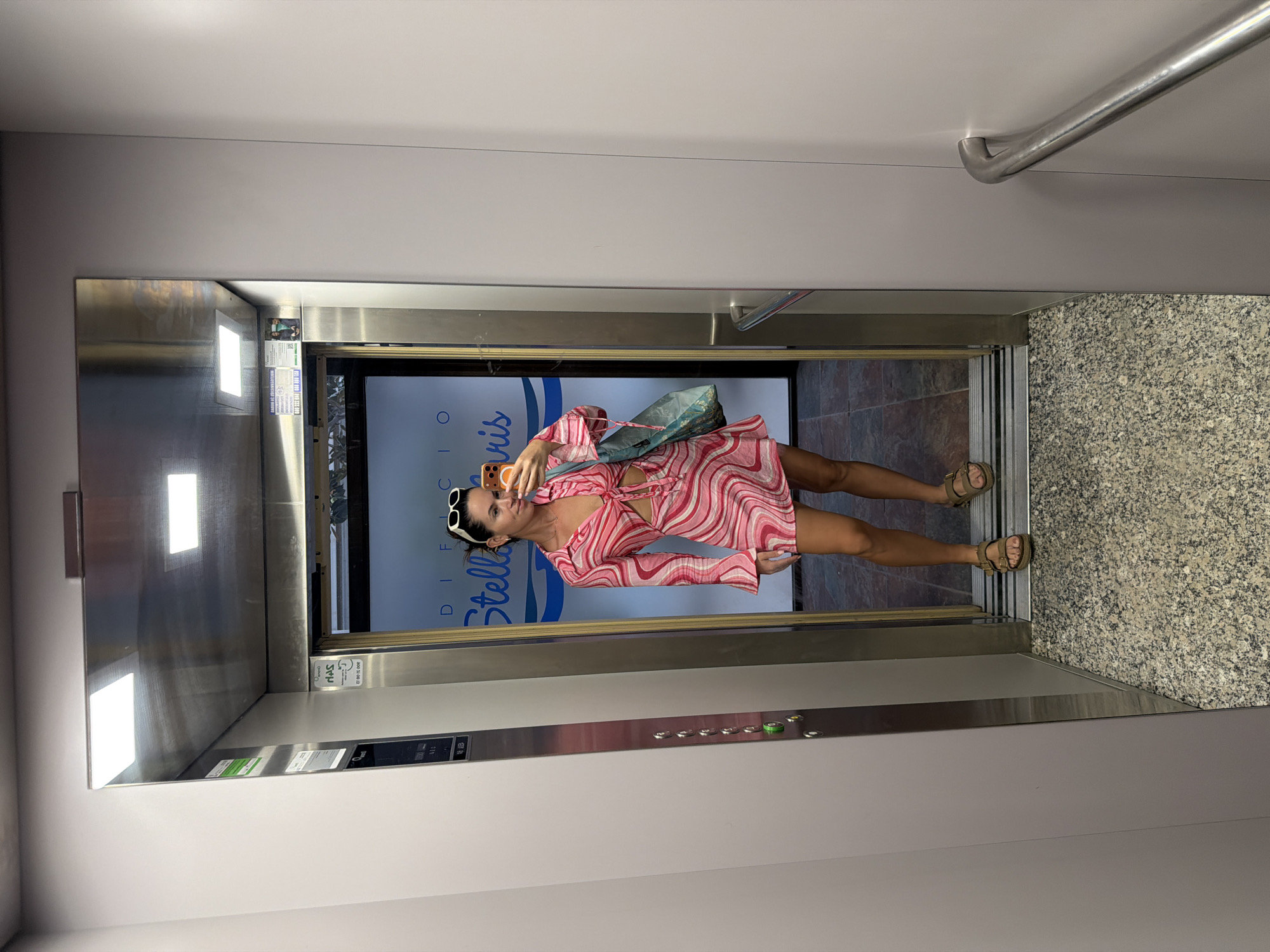 A young woman is standing in an elevator, taking a mirror selfie. She is wearing a pink and white swirled pattern dress with cutouts at the waist and bell sleeves. She has on brown sandals and white sunglasses pushed up on her head. She is holding an orange smartphone in her right hand. A blue and green patterned tote bag is slung over her left shoulder. The elevator interior is stainless steel with buttons on the left side. The floor of the elevator is a speckled gray material. The doors of the elevator are open, revealing a hallway with a blue sign that reads "DIRECCIO STELLA." The flooring in the hallway appears to be tiled in a dark red and brown pattern. The lighting in the elevator is bright and fluorescent. The woman appears to be in the city of Salou, Spain, as indicated by the sign.