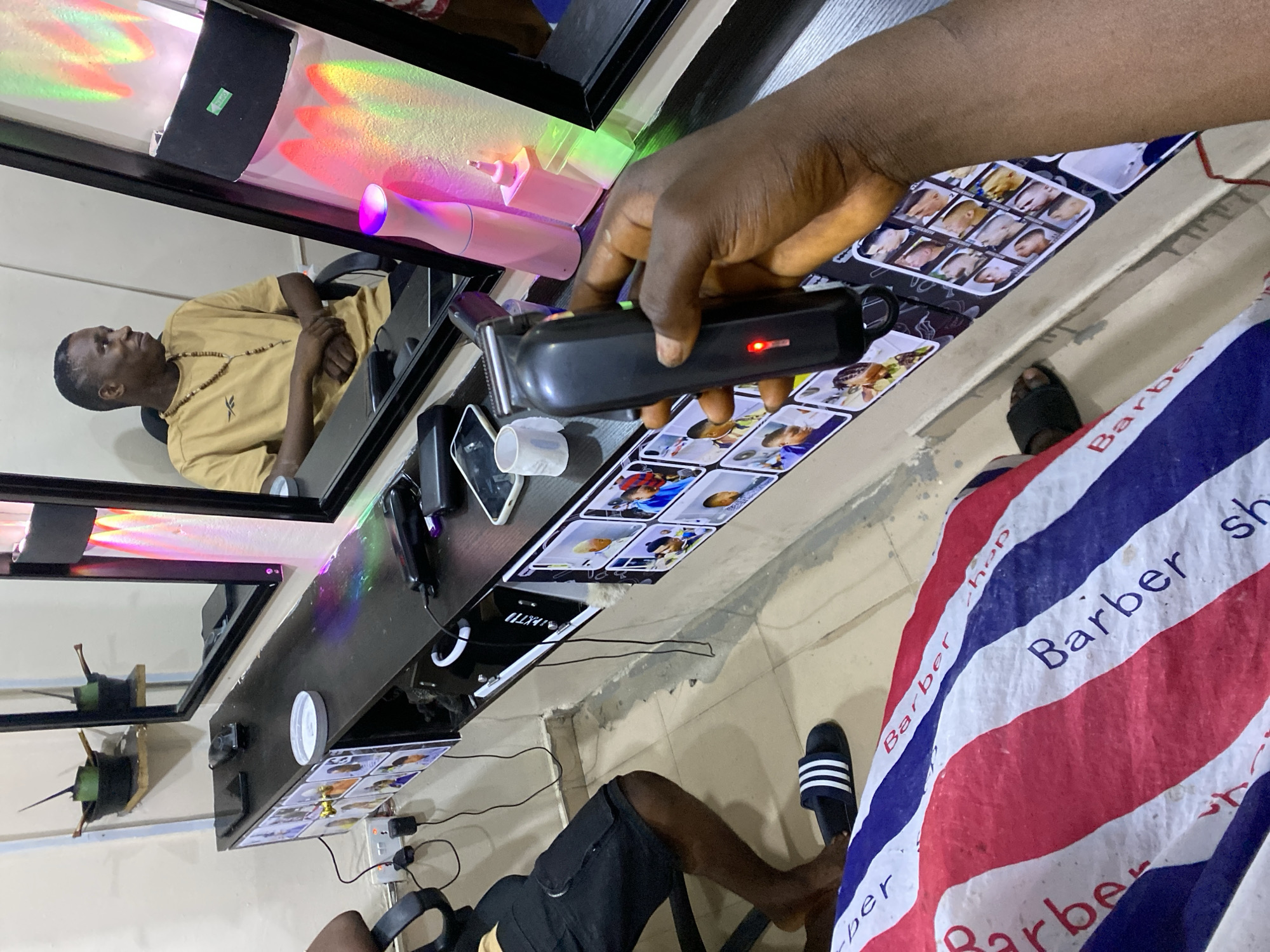 A photograph, rotated counterclockwise, depicts a scene within a barbershop in Ado, Nigeria. An African male with dark skin and short hair sits in a barber chair visible in the mirror and wearing a gold t-shirt and beaded necklace. The barber holds a black electric hair clipper with a red light illuminated near the bottom, over a counter showing sample hairstyles on picture tiles along the wall. The shop has a barber's cape with red, white, and blue stripes, with the word "Barber" repeated across the fabric, draped over the lower part of the image. A black slider sandal is visible beneath the cape. The barber counter is cluttered with objects, including small white containers, a phone, and various barber tools. A power strip and electrical cords are also visible under the counter. The environment is illuminated with colorful lighting, including a strip light above the mirror.