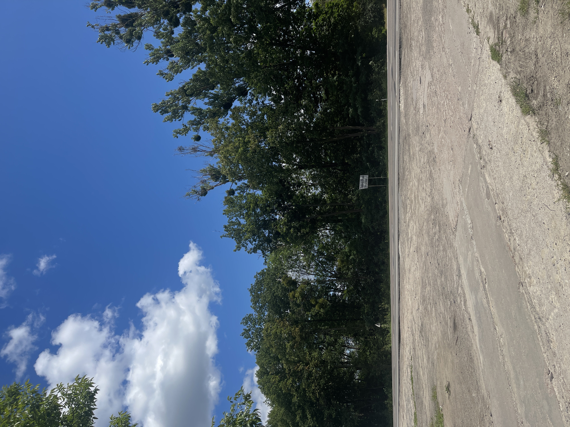 The image captures a roadside scene in Konopnytsia, Ukraine. In the foreground, a rough, unpaved area with patches of concrete and dirt dominates the lower portion of the frame. A paved road runs horizontally across the middle ground. Beyond the road, a dense line of trees forms a natural barrier. The sky above is a vibrant blue, with scattered white clouds adding texture. A white sign with text in Ukrainian and English, "Зимна Вода / Zymna Voda," is visible to the left of the trees, indicating a nearby location. The overall environment suggests a rural or semi-rural setting. No people or vehicles are present in the image.