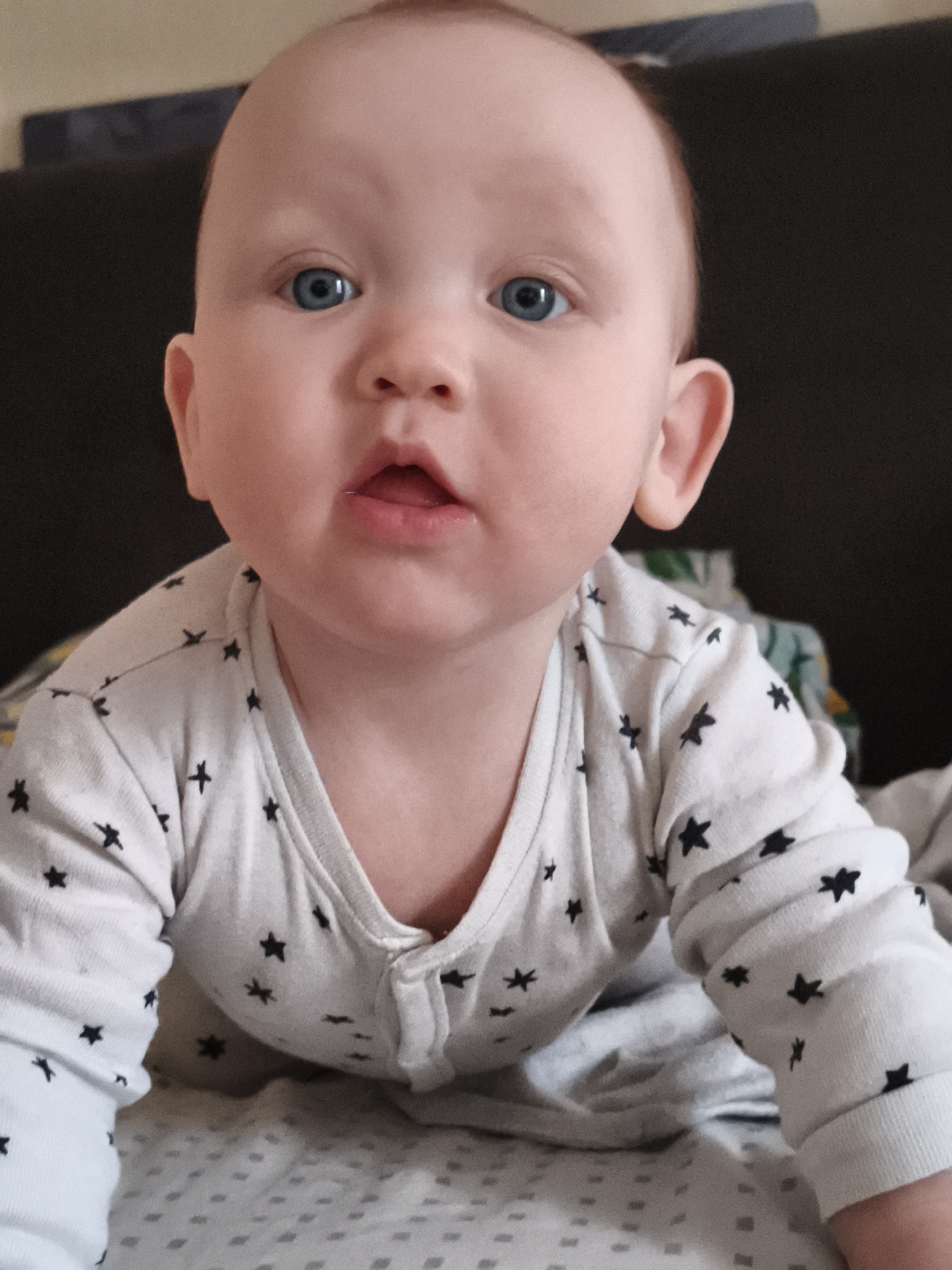 An infant is depicted in a close-up, chest-up view, appearing to be lying prone and looking directly into the camera. The infant has fair skin, prominent blue eyes, and minimal light-colored hair. Its mouth is slightly open, revealing the tongue. The infant wears a white, long-sleeved garment patterned with numerous small black stars. This garment has a V-neckline and three small white buttons down the front. The immediate surface beneath the infant is a light grey fabric with a repetitive pattern of small dark squares. The background is out of focus, consisting primarily of a dark, uniform surface, likely upholstery, behind the infant's head and shoulders. A small, indistinct area of lighter, possibly green and white patterned fabric is visible to the upper right in the background. The specific location within the city of Chaikovsky, Russia, cannot be determined from the visual information presented in this image.