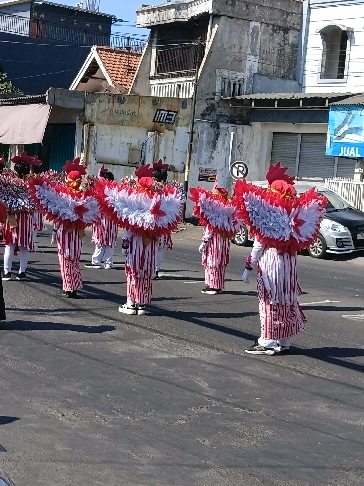In Wadungasri, Indonesia, on a sunny day, a parade is underway featuring participants in striking red and white costumes. These individuals are adorned with elaborate headdresses and large, wing-like embellishments on their backs, predominantly in red and white. They are dressed in striped trousers and shirts, with some wearing white socks and sneakers. The parade is taking place on an asphalt road, with buildings and a parked silver car visible in the background. The buildings display various architectural styles, some appearing weathered and aged. Signs and electrical wires are also present in the urban environment. The scene captures a festive moment in the community, with the participants likely engaged in a cultural or celebratory event.