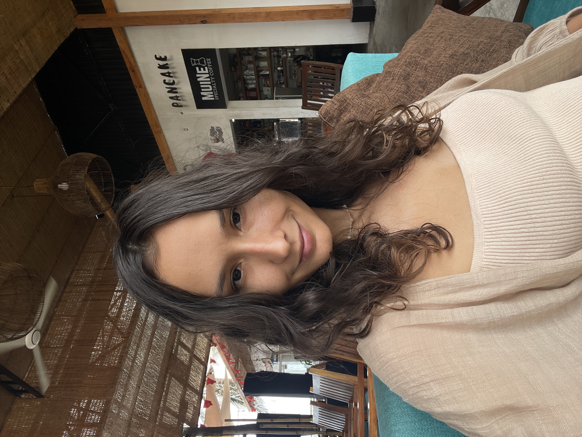 A medium shot captures a young woman with long, dark, wavy hair and brown eyes, smiling gently at the camera. She is seated on a turquoise upholstered bench, with a brown textured pillow to her right. She wears a light beige ribbed tank top and a matching, open-front cardigan. Behind her, an open-air establishment features wooden chairs and tables, a thatched roof overhead, and a sign that reads "MUINE SPECIALTY COFFEE" and "PANCAKE". The location appears to be an outdoor cafe in Phu Thuy Ward, Vietnam, suggesting a tropical or coastal setting with ambient lighting and natural materials used in the architecture.
