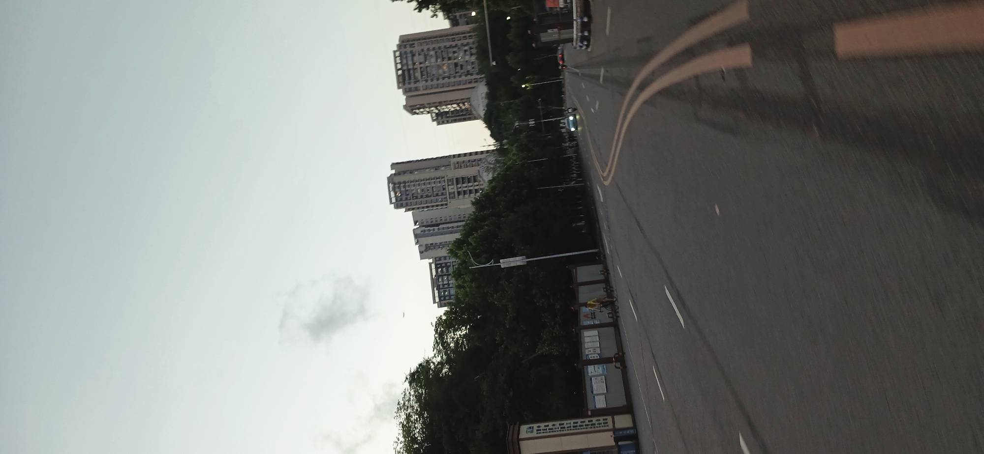 The image shows a street in Jiulongpo District, China. High-rise apartment buildings line the left side of the street behind a row of trees. A double yellow line runs down the middle of the asphalt road, and there are occasional white stripes marking lanes. A bus stop shelter is visible on the left side of the road, and a person is near it. The sky is overcast. Several vehicles can be seen in the distance.