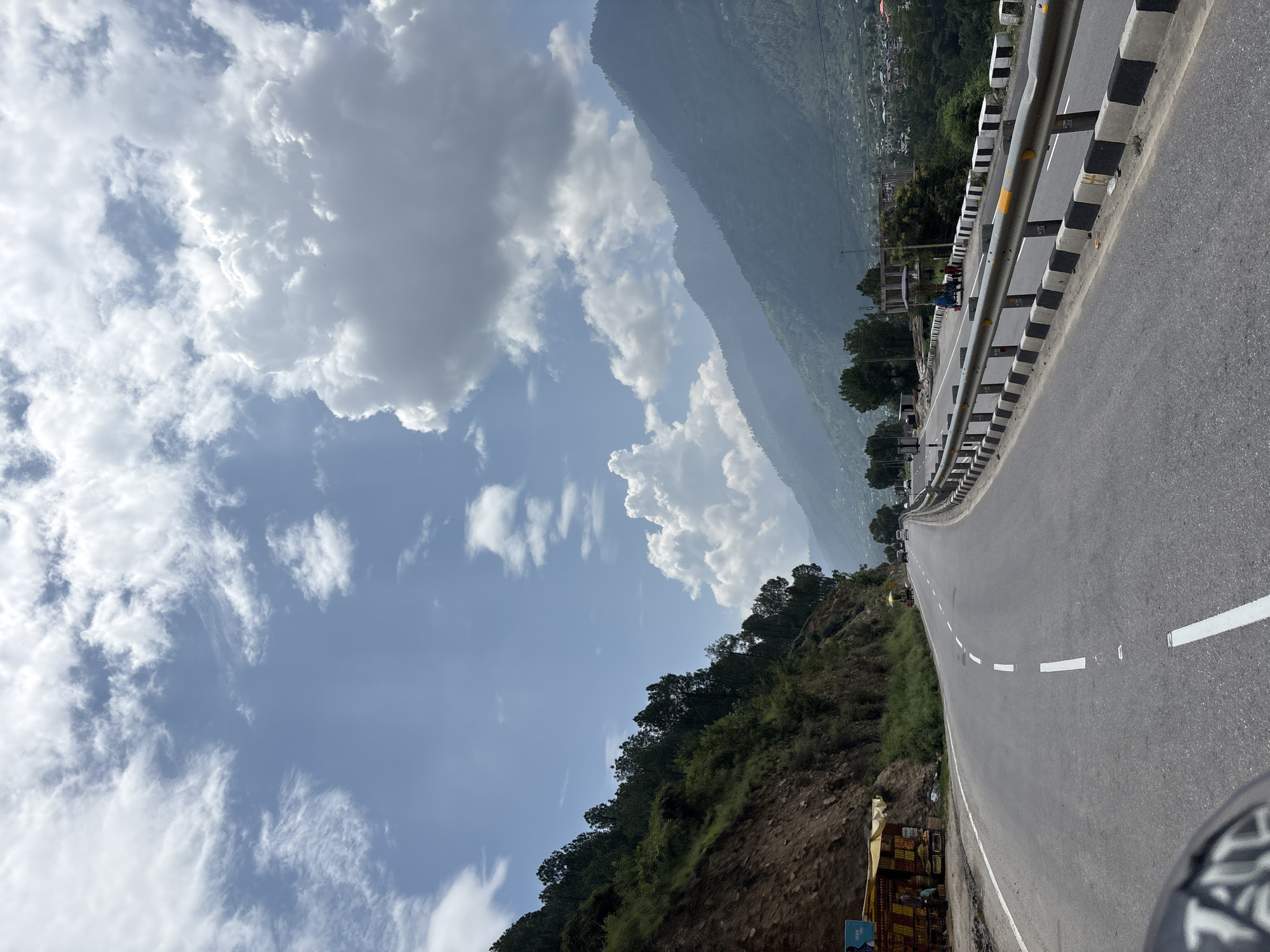 A wide, curved asphalt road cuts through a mountainous landscape. The road is bordered by a concrete barrier with black and white stripes. On the left, a steep, green, tree-covered mountainside rises. On the right, the road slopes down into a valley with a town nestled in the distance. The sky is a bright blue with fluffy white clouds. A few cars and a cart are visible on the road and roadside. A small roadside stall with colorful goods is on the left. The scene appears to be in Pirdi, India, based on the characteristic terrain and roadside structures.