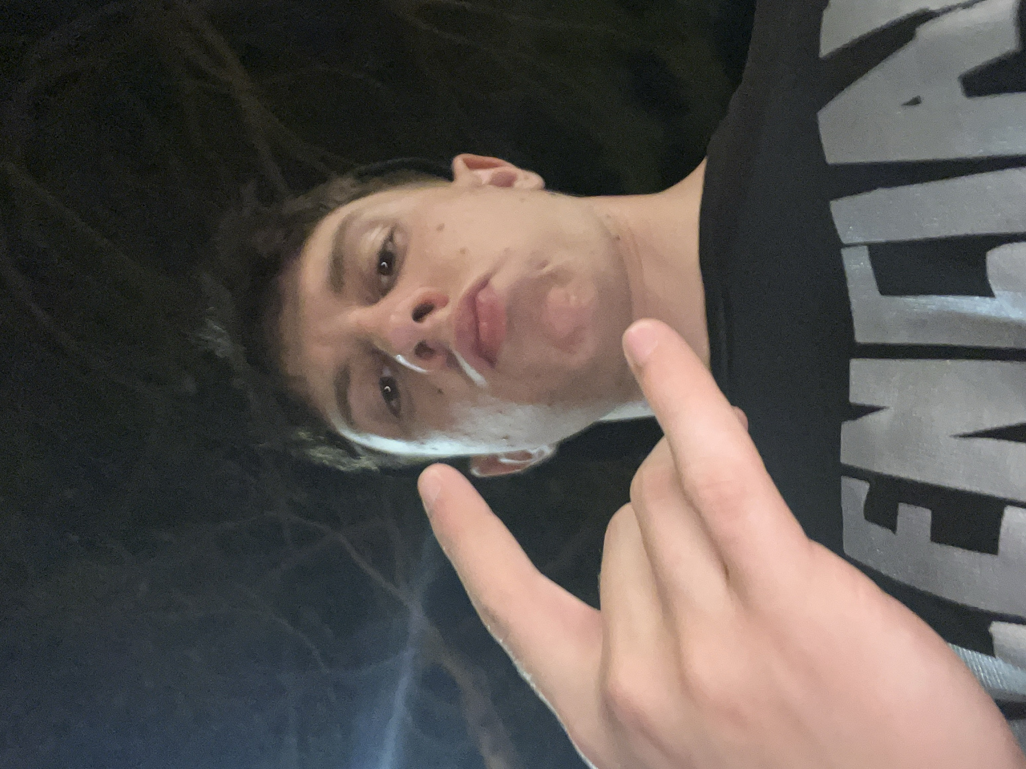 A person with light skin and short, dark hair is positioned at an acute angle to the camera, face-up, with a finger held up close to the lens. The individual's lips are pursed. The person is wearing a black shirt with a large, light gray graphic. The background shows dark tones suggesting an evening or nighttime setting with vague, undefined shapes. The location in the city of Jesolo, Italy, is not discernible from the visual information provided.