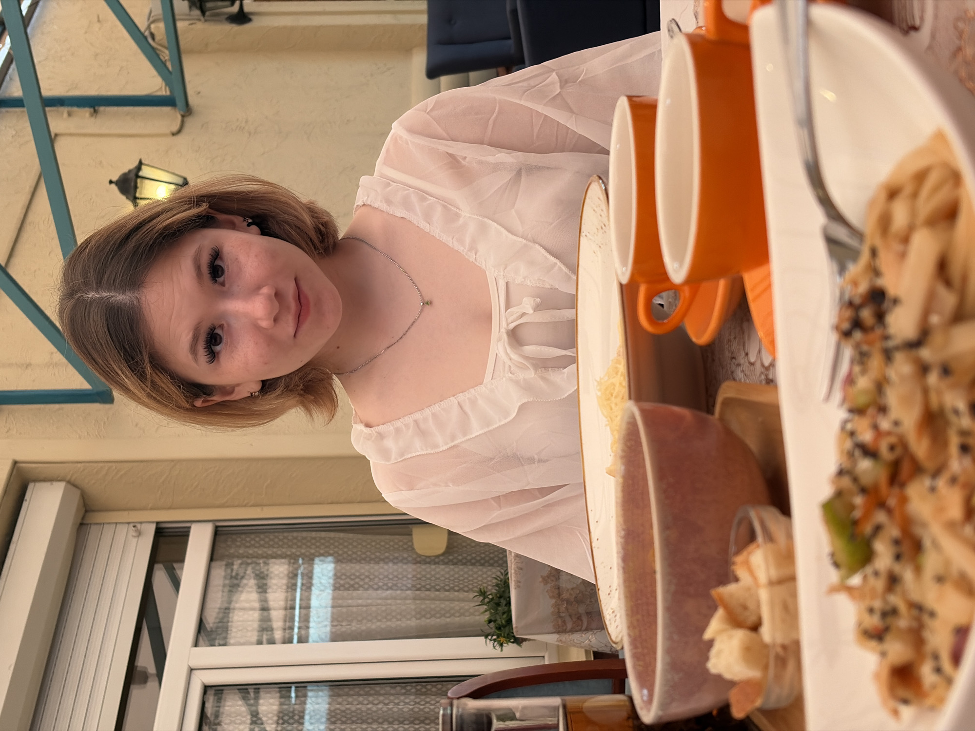 In an outdoor setting, a young woman with short brown hair and expressive eyes smiles at the camera. She is wearing a sheer white blouse with ruffled detailing and a delicate silver necklace. The image is oriented sideways, giving a unique perspective. In the background, a warm-toned wall is visible, along with an outdoor lamp that suggests it could be evening or a dimly lit area.
To the right of the woman, a table is set with several orange mugs and a white plate filled with what appears to be a salad or pasta dish, garnished with black sesame seeds and vegetables. A pinkish-brown bowl, partially filled with small pieces of bread, sits next to the plate. The setting appears to be a cafe or restaurant, with a glimpse of window blinds and foliage in the background, hinting at a potentially pleasant, perhaps warm, weather. The overall mood is relaxed and friendly, with the woman's smile conveying a sense of happiness.