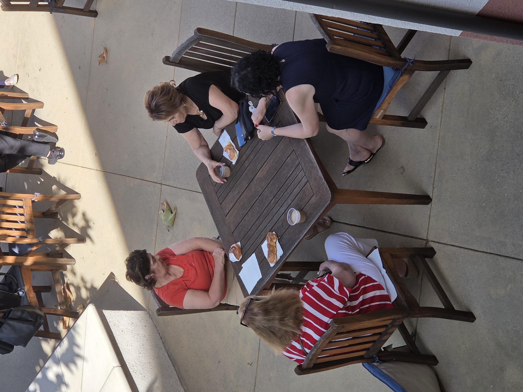 Four individuals are seated at a wooden outdoor table, likely at a cafe or restaurant, in Glendale, United States of America. The scene is outdoors with concrete flooring and dappled sunlight indicating a sunny day. Two of the individuals are women seated opposite each other at the table. One woman, wearing a red top, is looking down at the table. The other woman, wearing a black top, is holding a small plastic cup. To the left of the table, another woman in a red and white striped shirt is seated and looking at her phone. Opposite her, a woman in a dark blue dress is also seated, looking towards the table. On the table are two pastries, napkins, a small plastic cup, and two cell phones. Several wooden chairs surround the table, and a large tote bag is visible on the floor next to one of the chairs. Leaves are scattered on the ground.